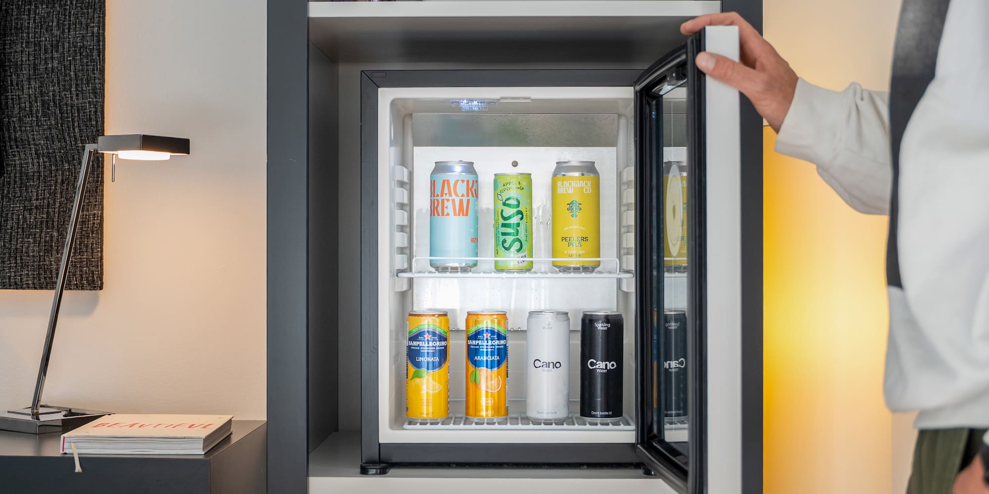 Person opening a hotel minibar stocked with various canned drinks.