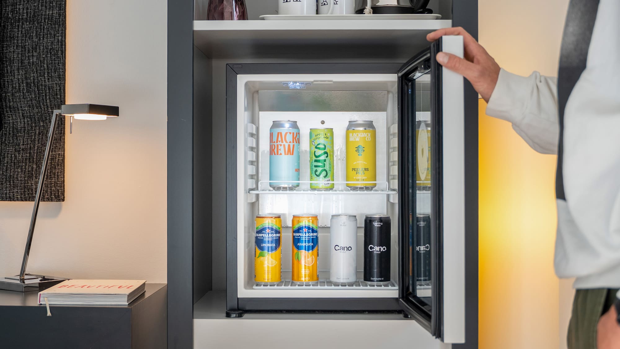 Person opening a hotel minibar filled with various canned sodas, water, and beer.