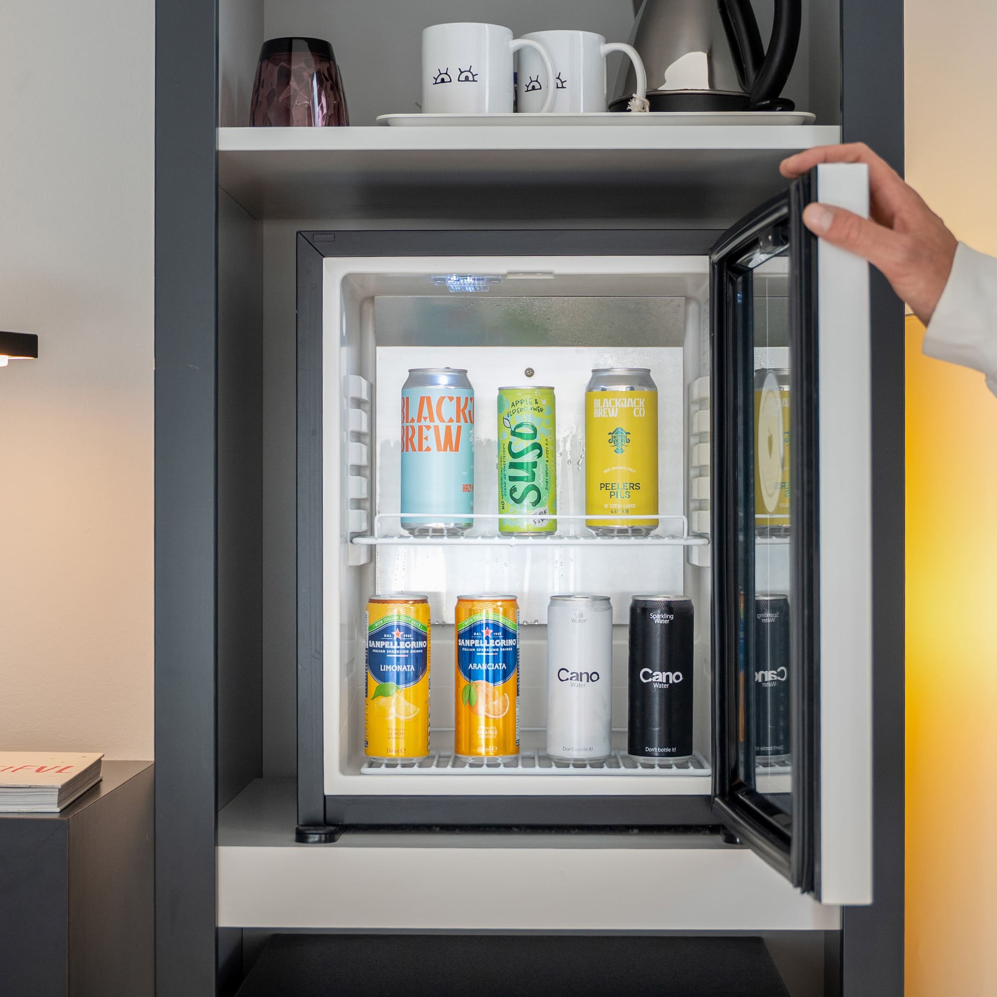 Person opening a hotel minibar stocked with various canned drinks.