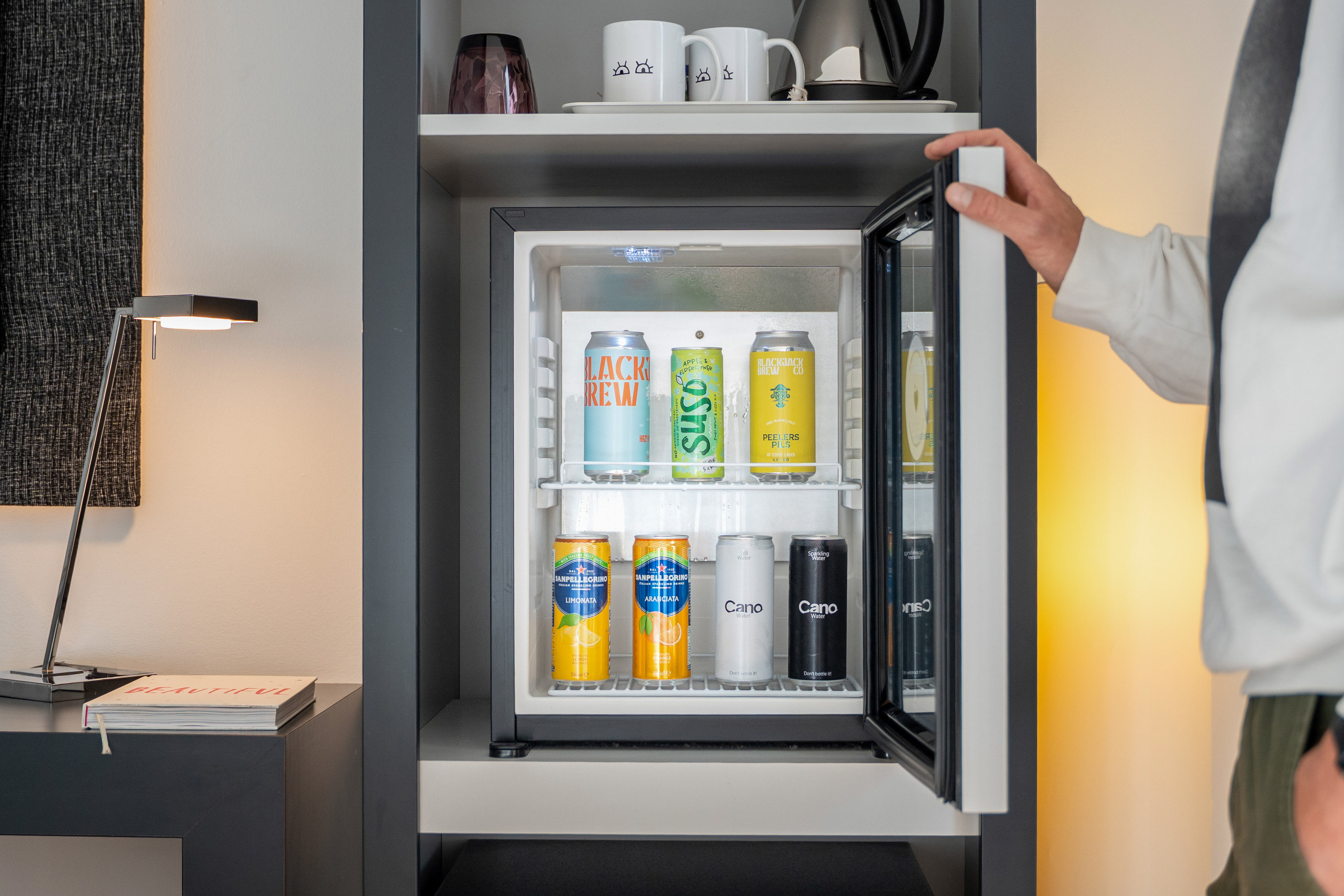 Person opening a hotel minibar stocked with various canned drinks.