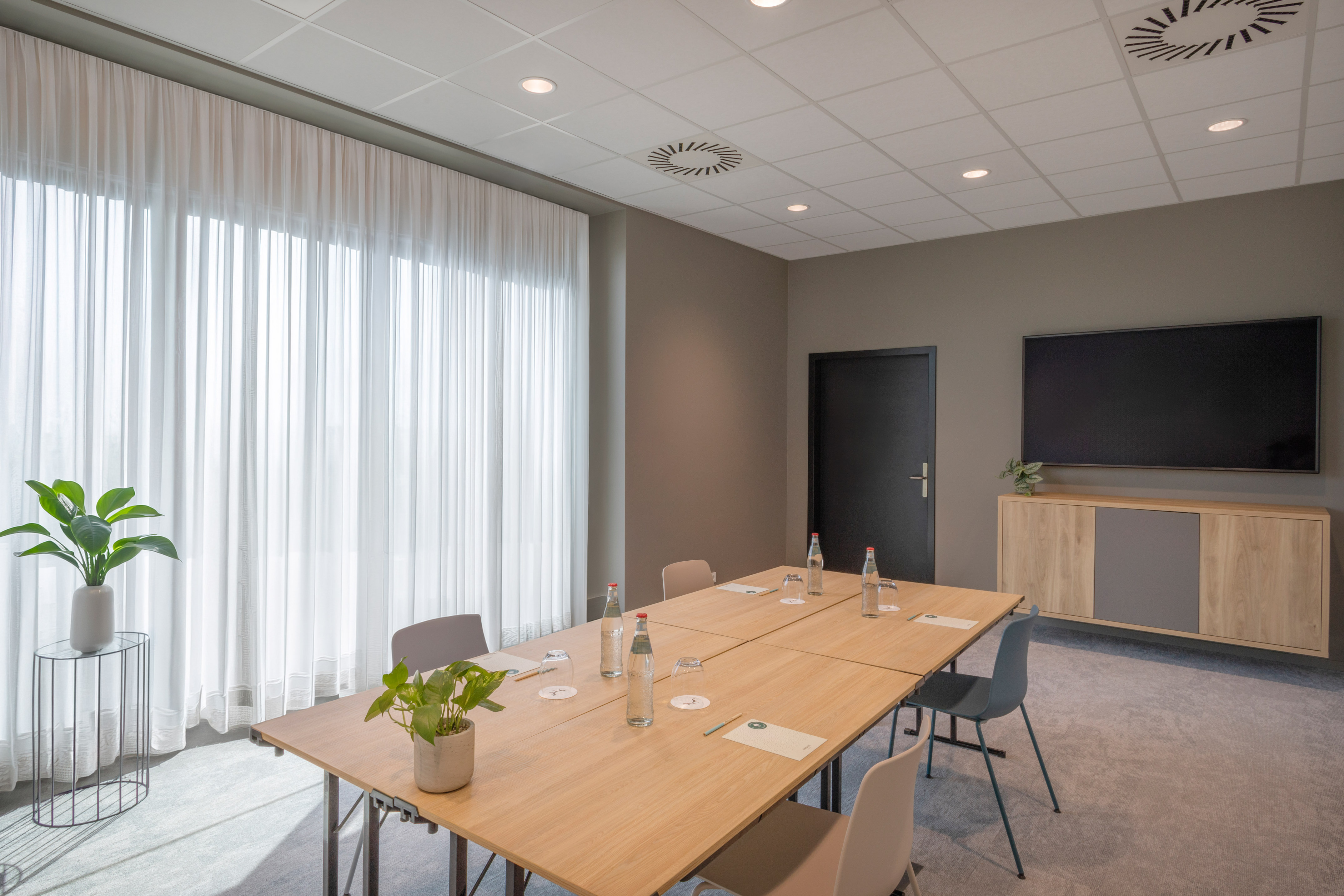 Modern meeting room with long table, TV, sheer curtains, and natural light.