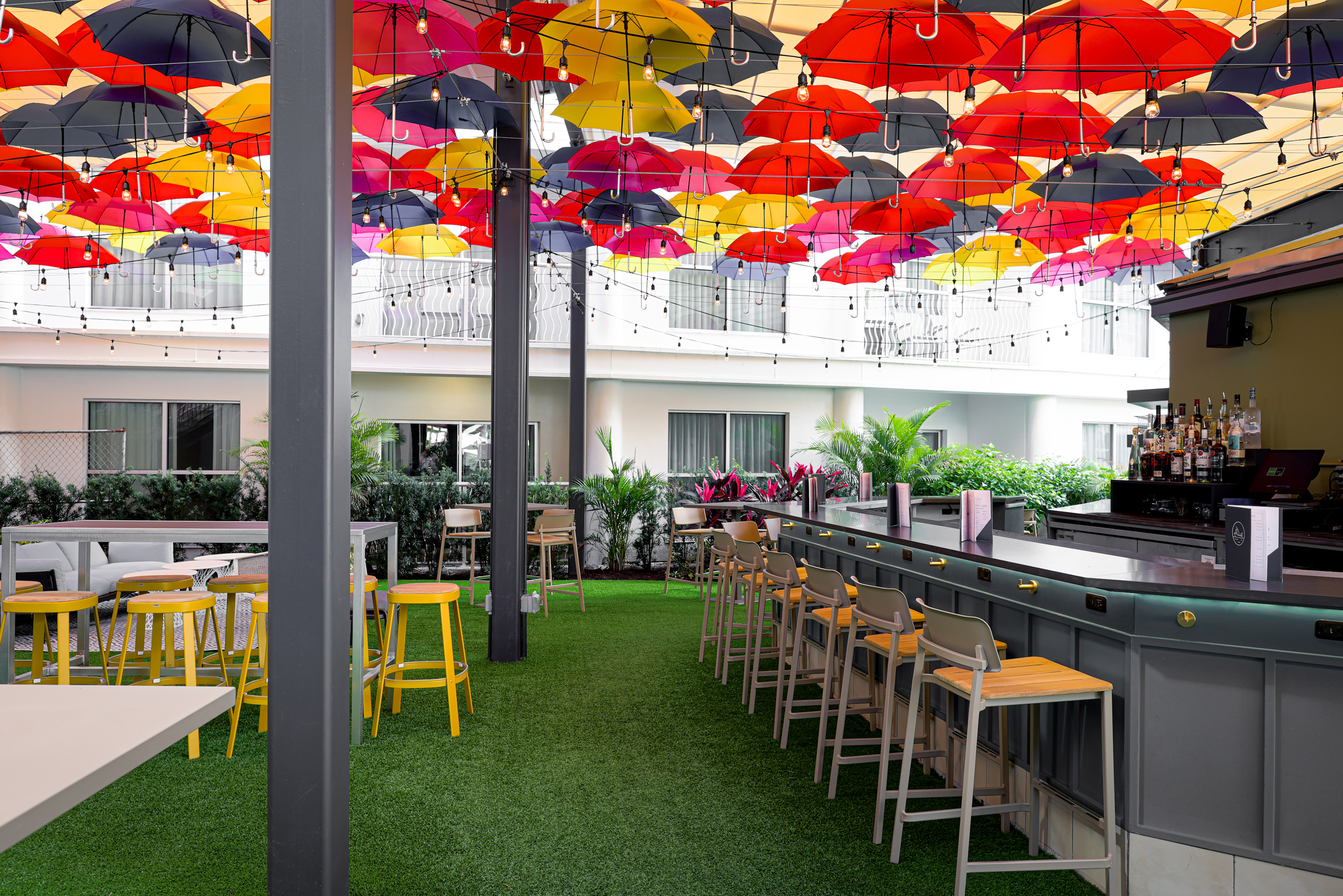 Vibrant outdoor bar with colorful umbrellas, string lights, and green turf.