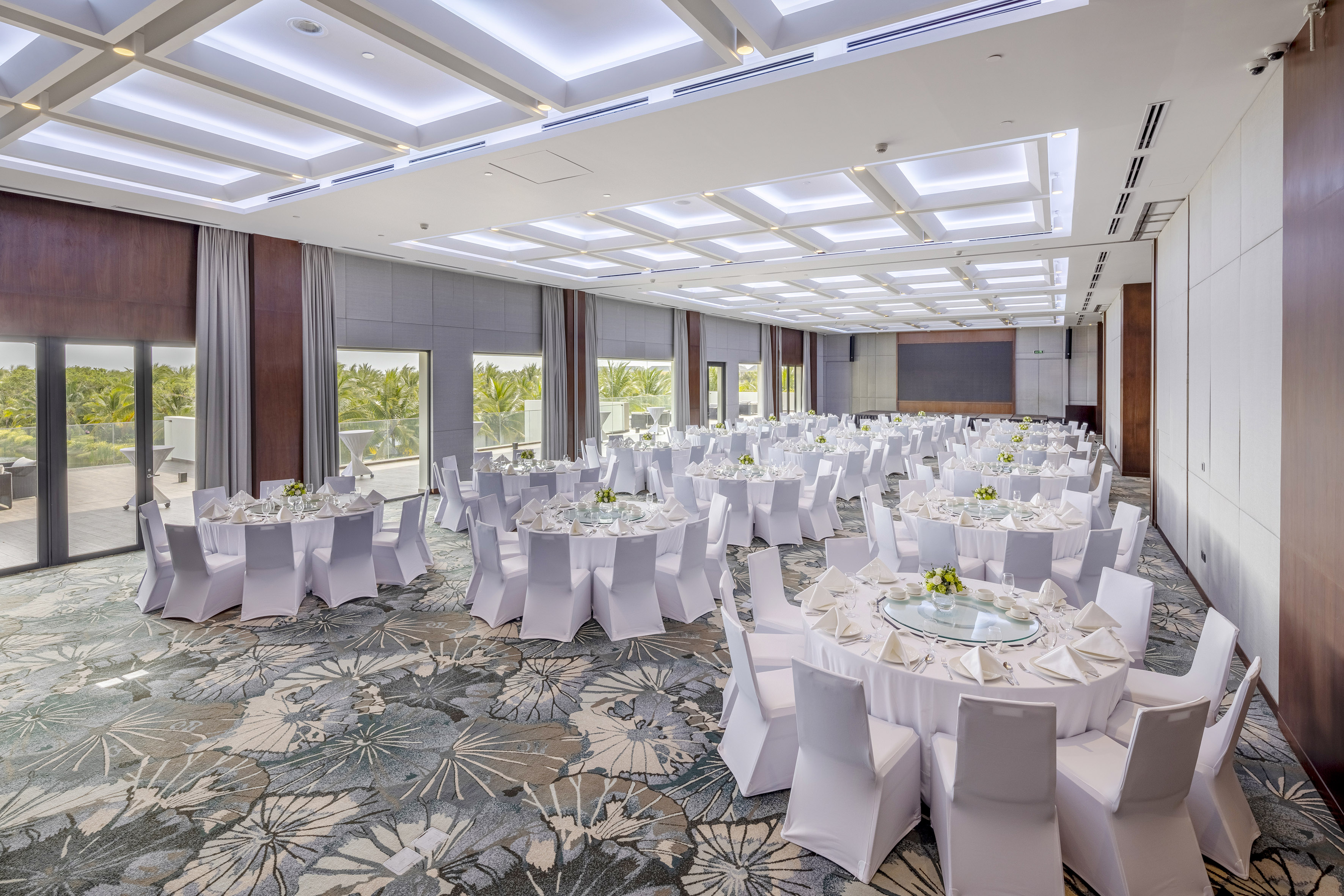 Modern hotel ballroom with white round tables, natural light, and tropical views.