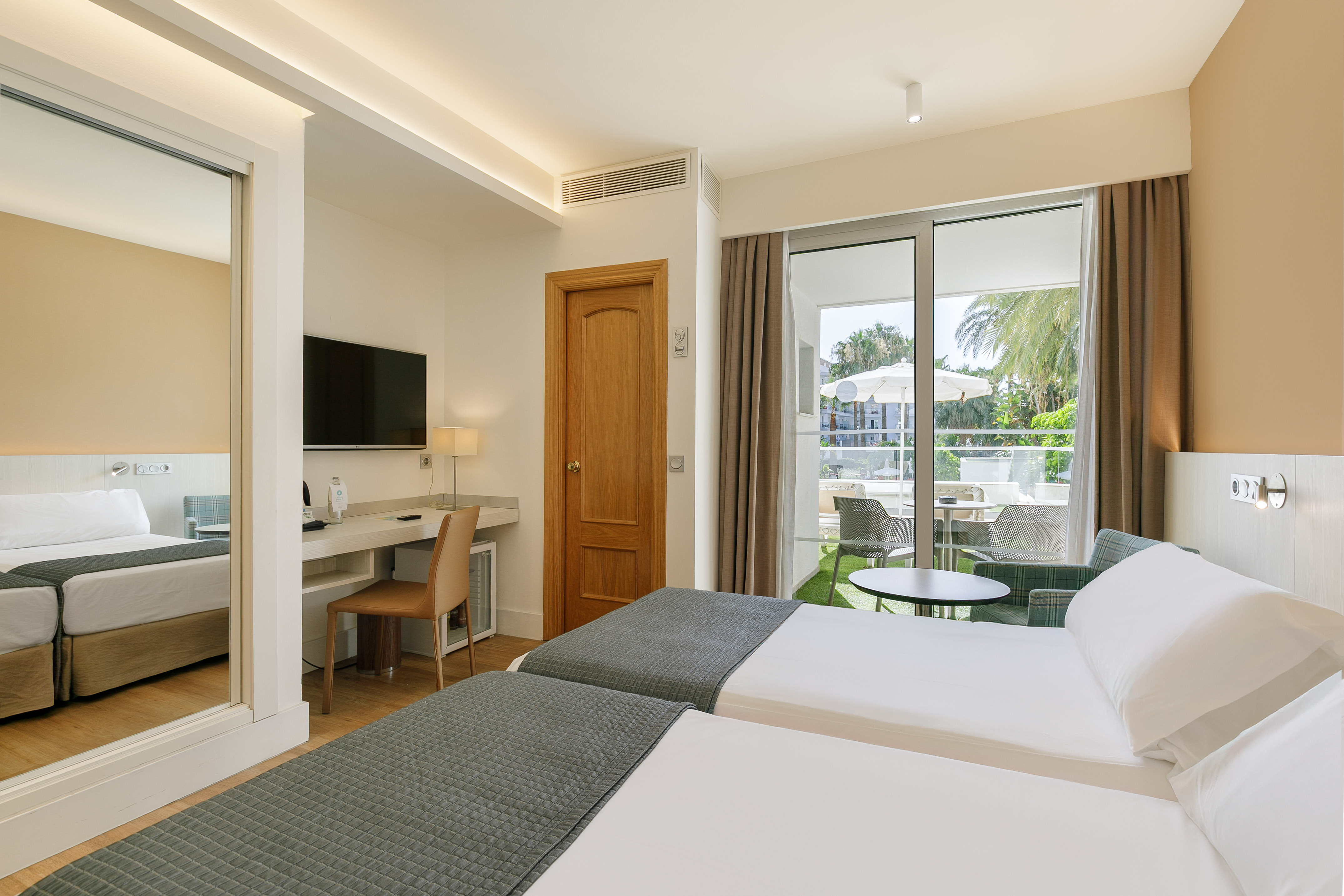 Modern hotel twin room with balcony, desk, TV, and tropical view.