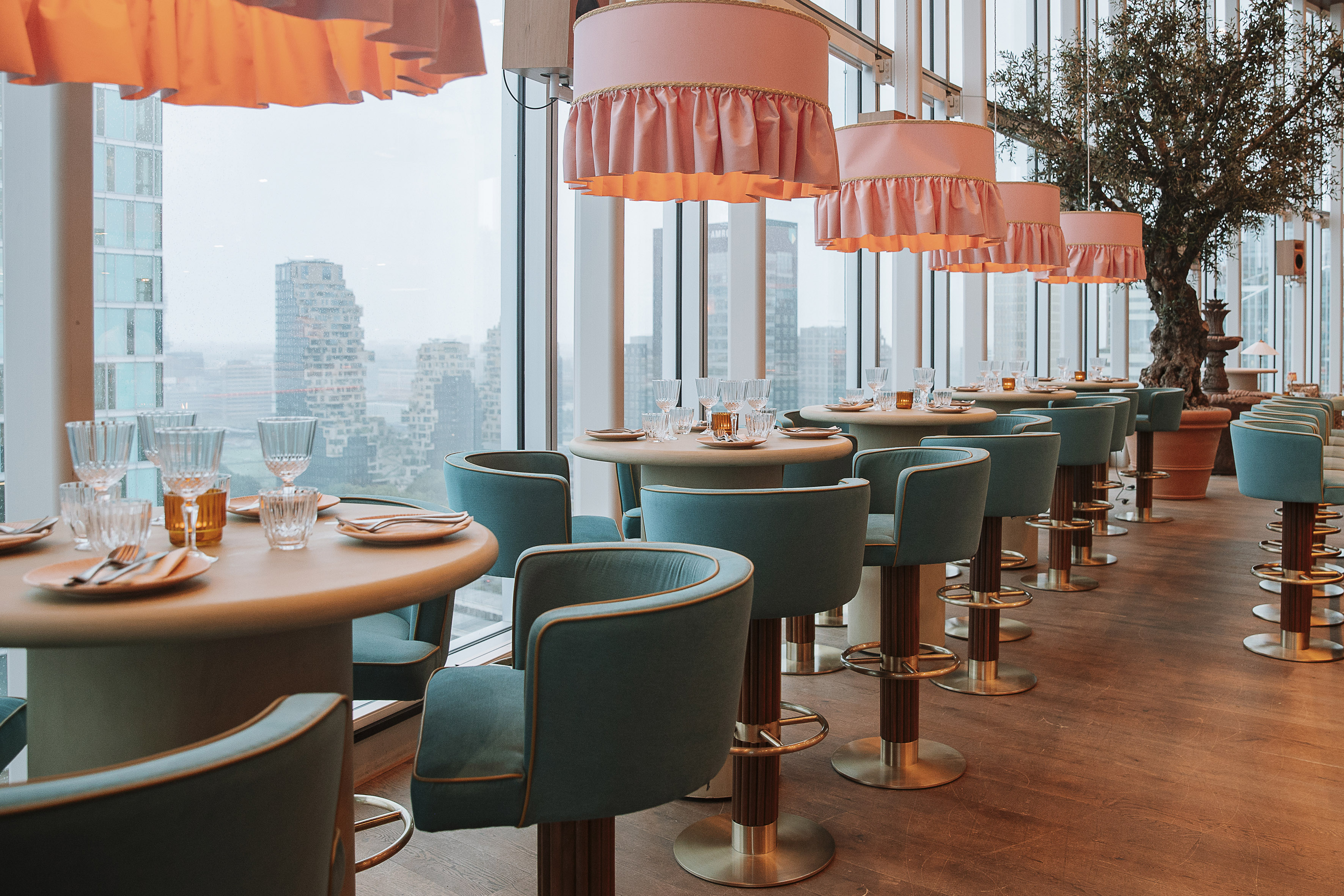 Elegant high-rise restaurant with city view, pink lamps, and teal dining chairs.
