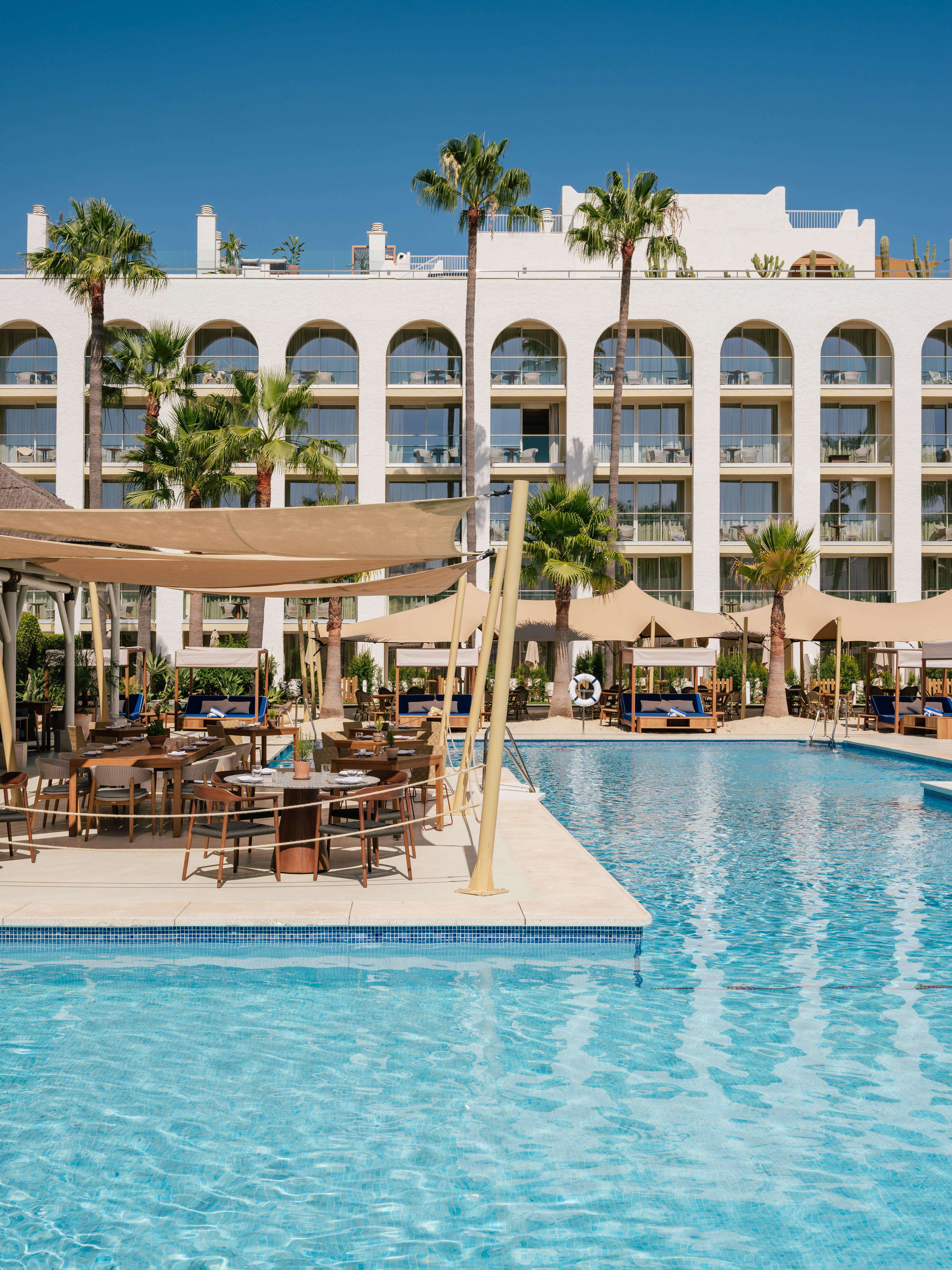 Hotel resort pool, outdoor dining, white arched building, palm trees.