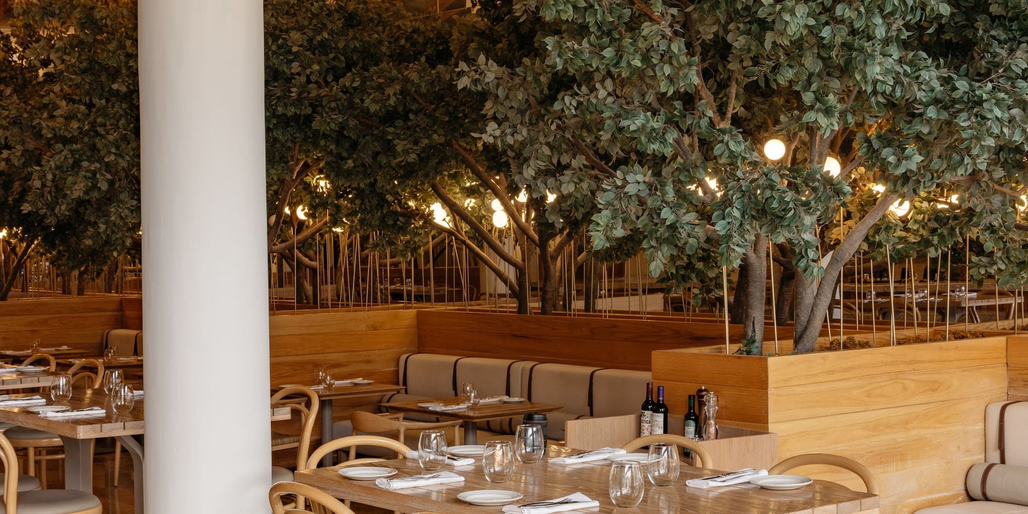 Restaurant dining room with wooden tables, rattan lamps, and indoor trees.