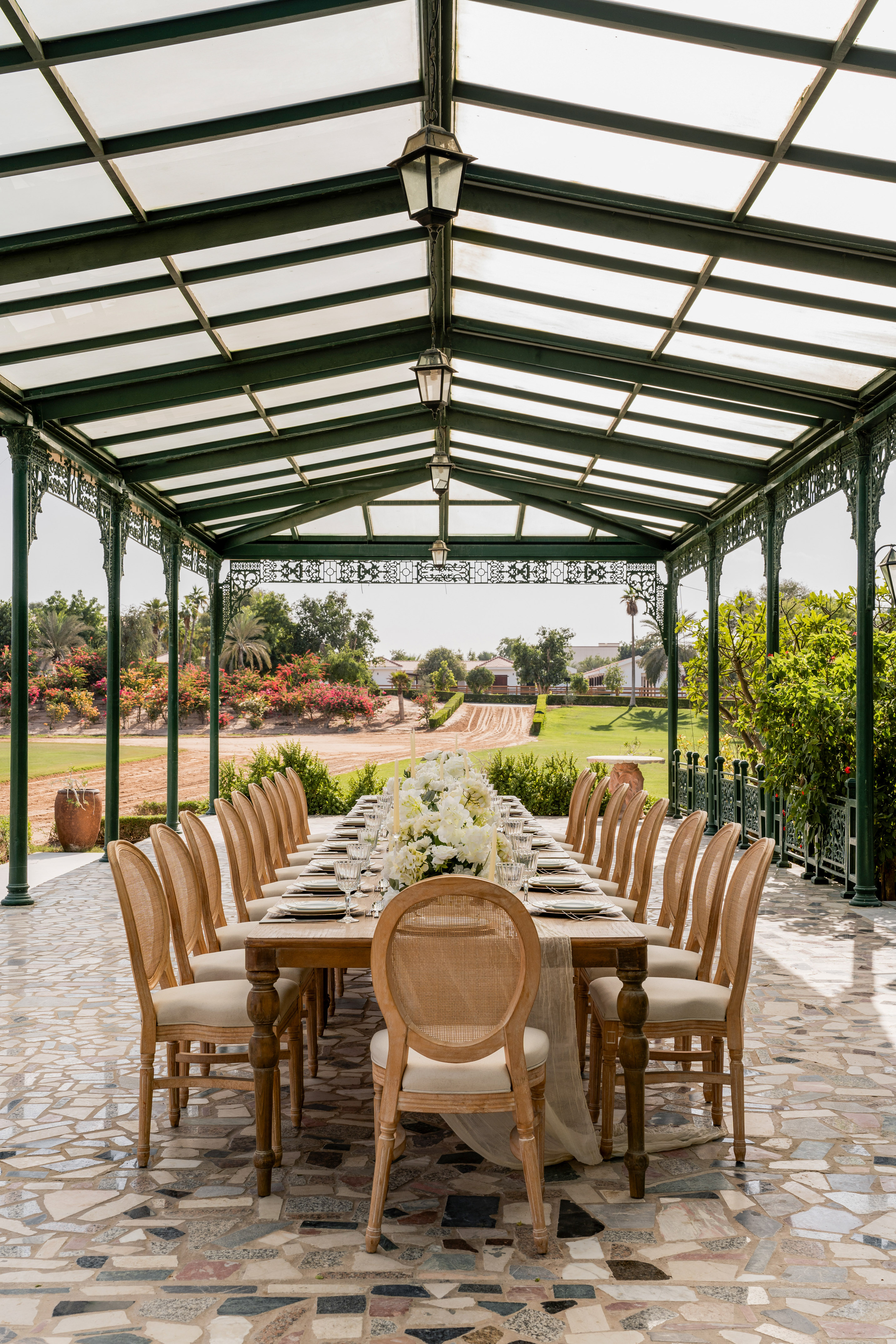 Outdoor dining table with white floral centerpiece under a green pergola.
