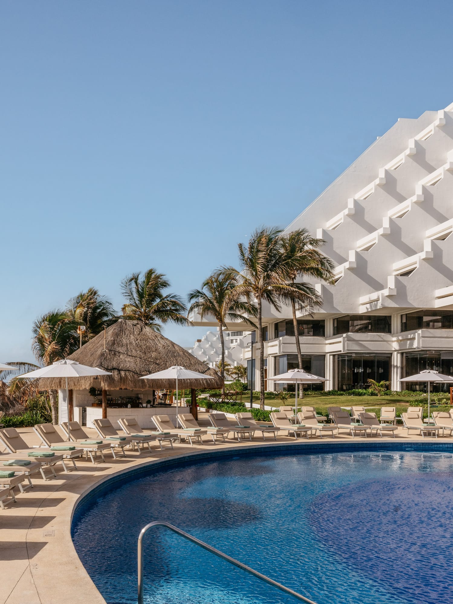 Tropical resort pool, white hotel, palm trees, lounge chairs, and ocean.