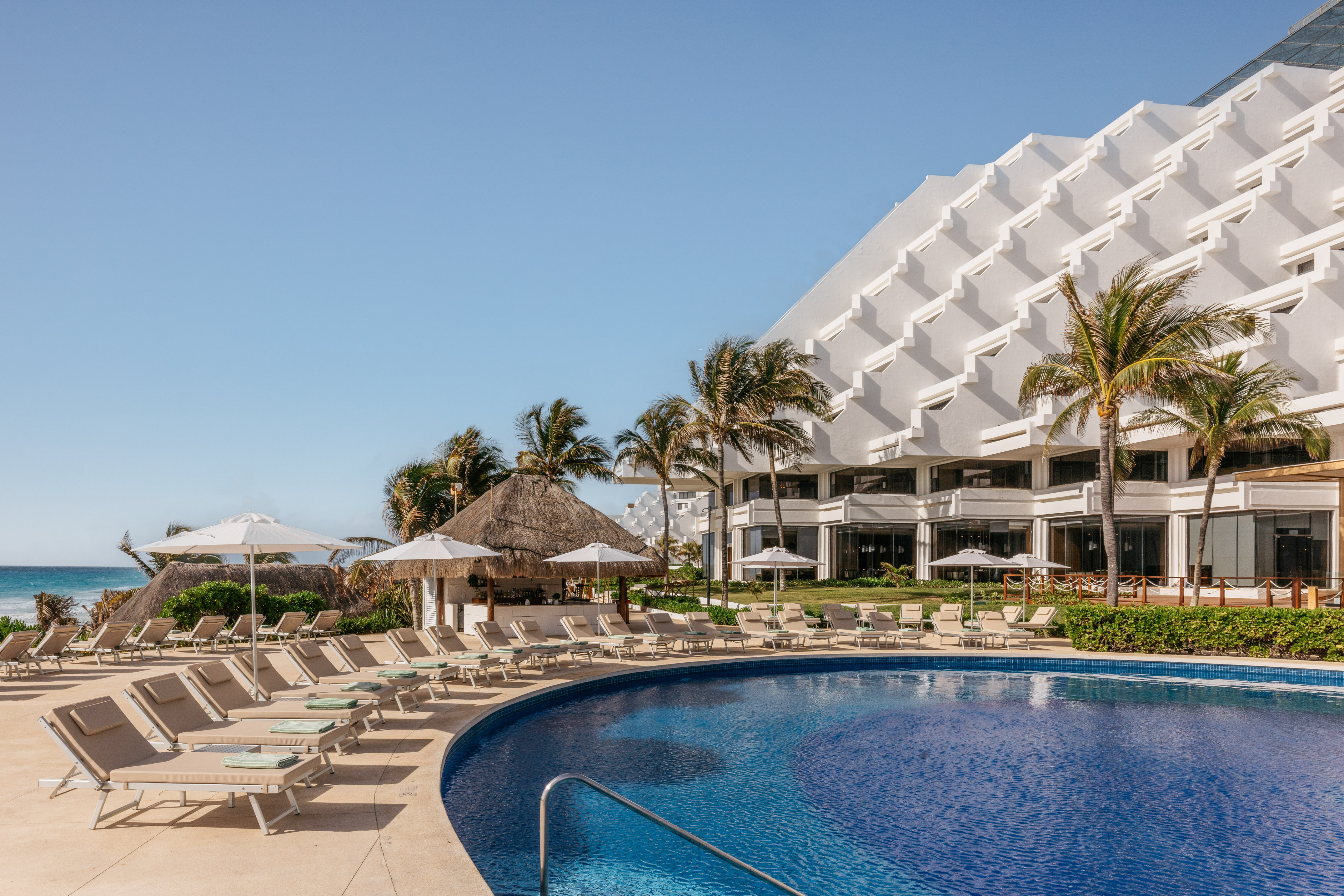 Tropical resort pool, white hotel, palm trees, lounge chairs, and ocean.