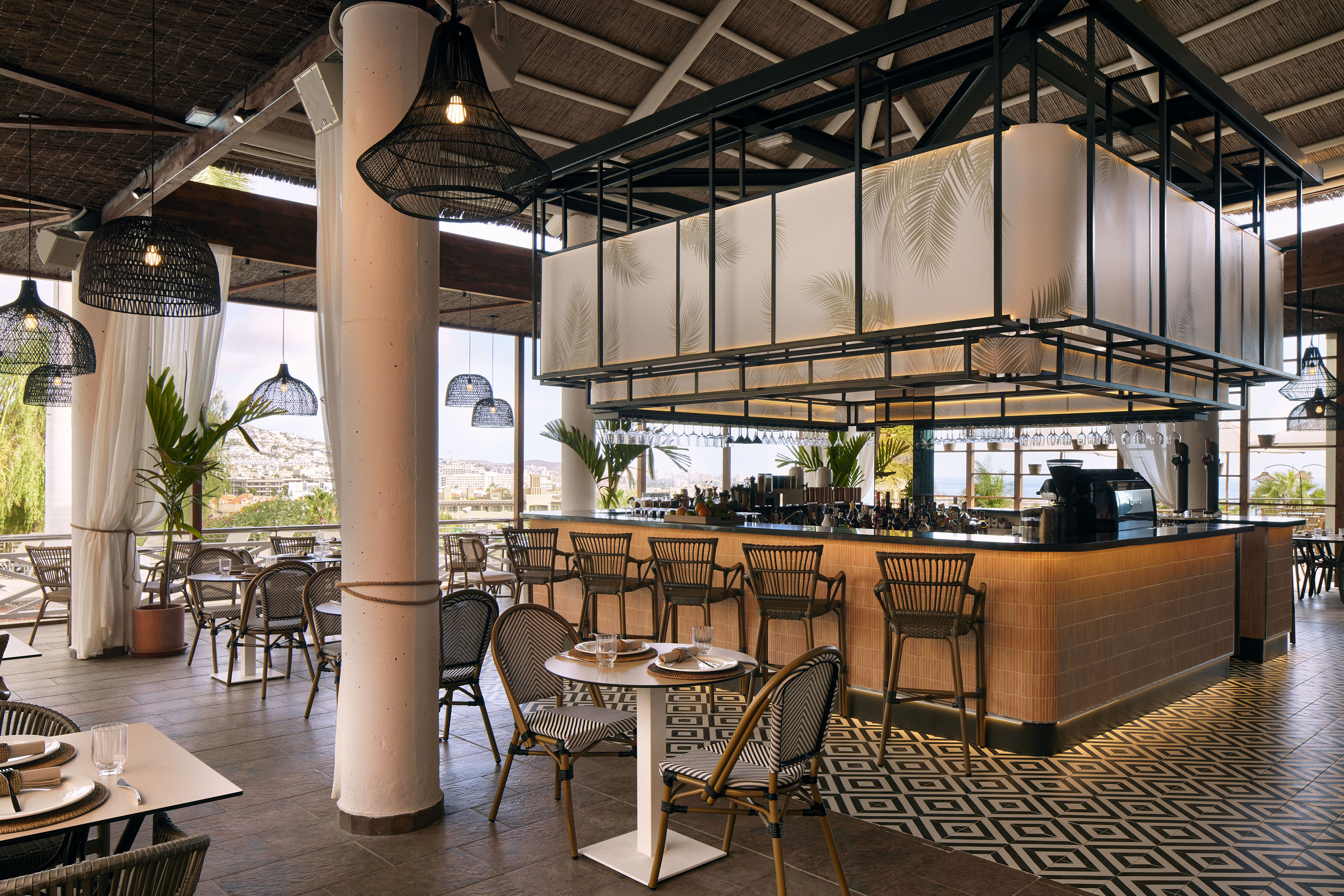 Stylish tropical resort restaurant bar with woven pendants, patterned floor, and city views.