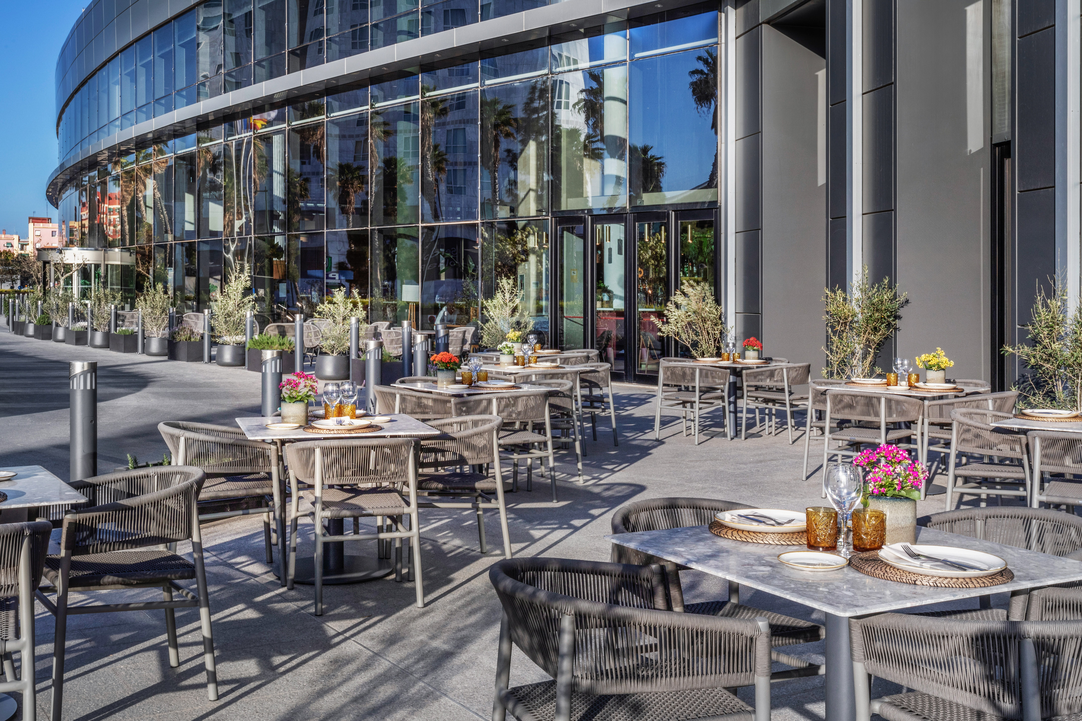 Modern outdoor restaurant terrace with glass facade, set tables, and plants.