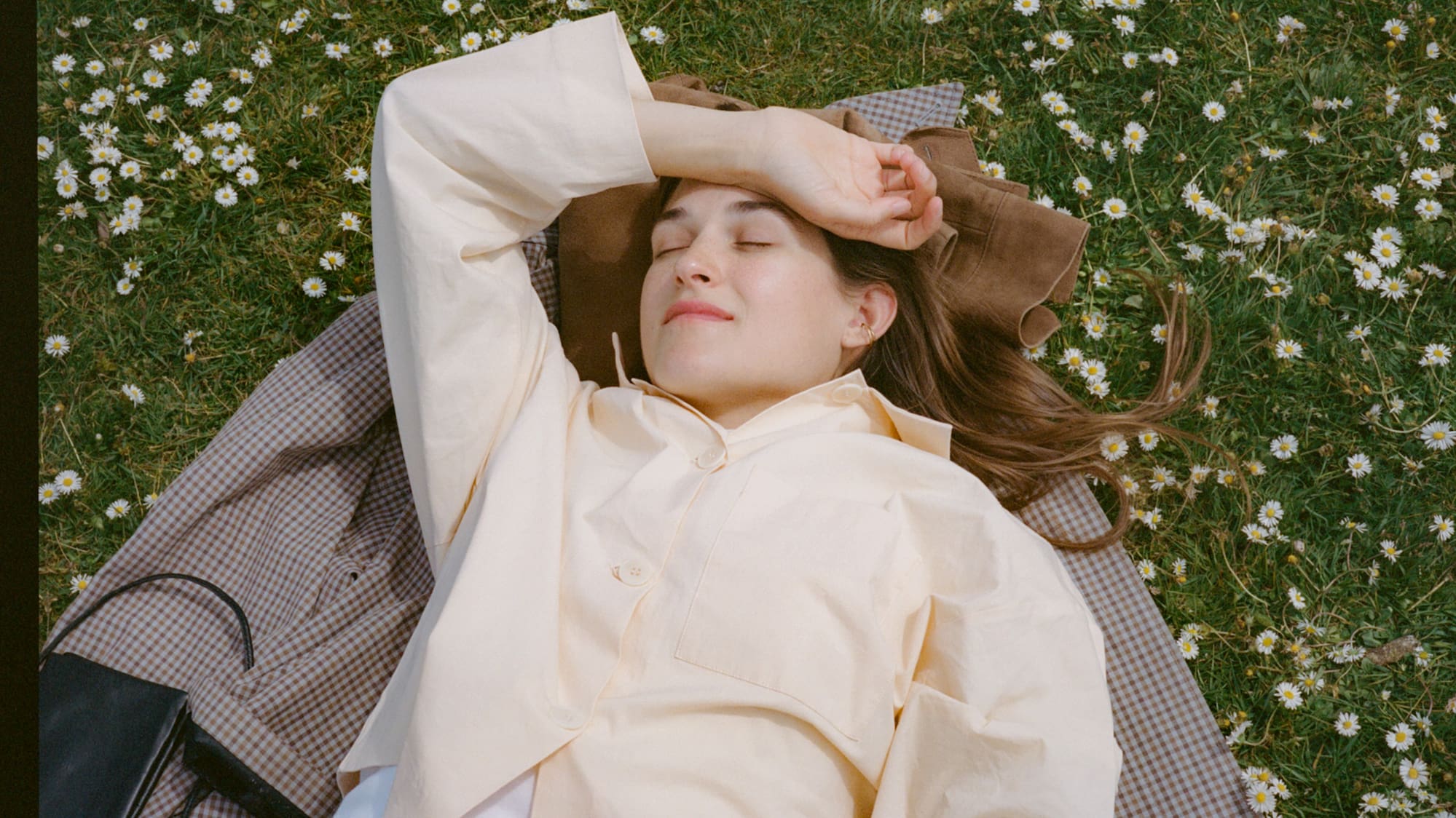 Woman napping on plaid blanket in daisy-filled grass with book.
