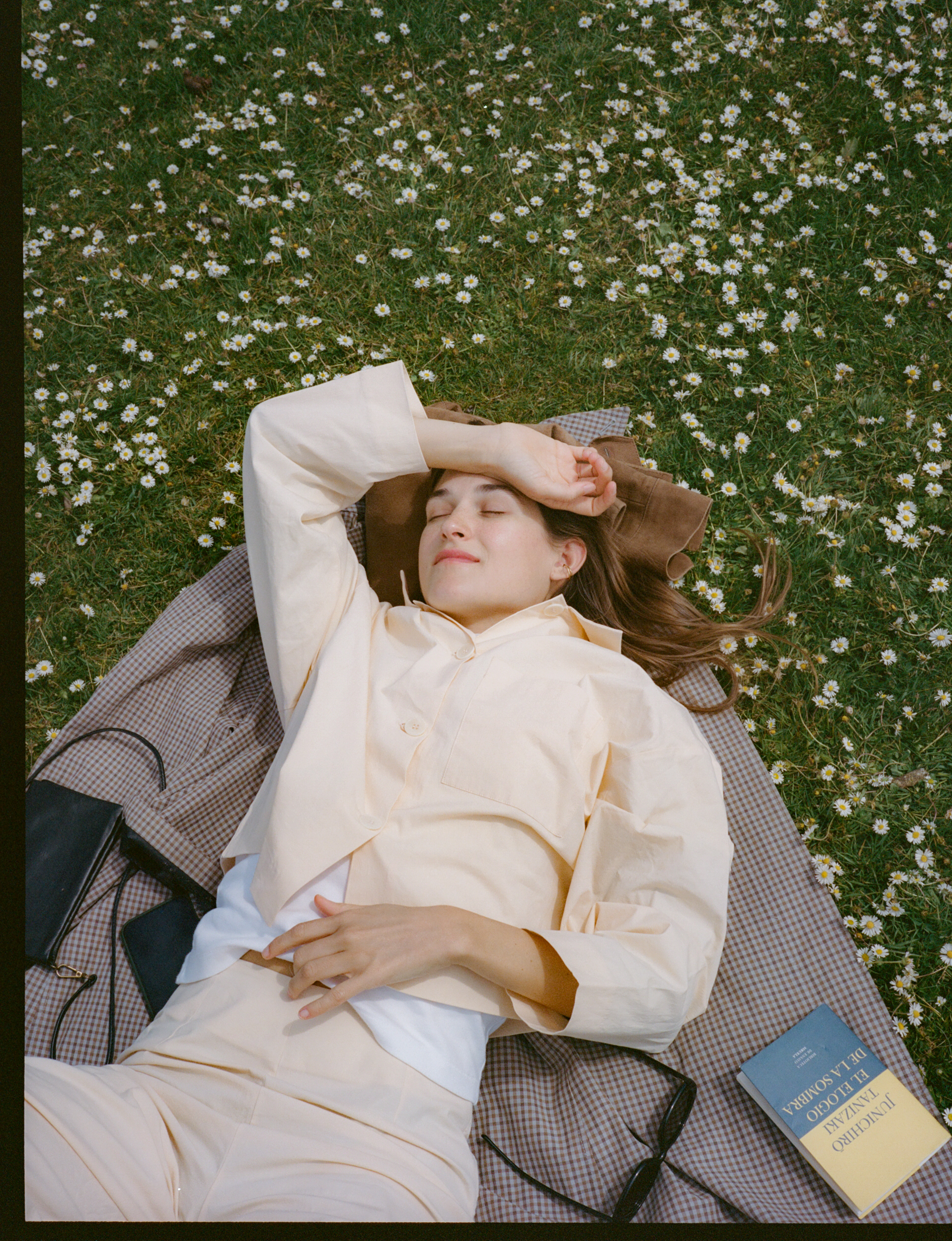 Woman napping on plaid blanket in daisy-filled grass with book.