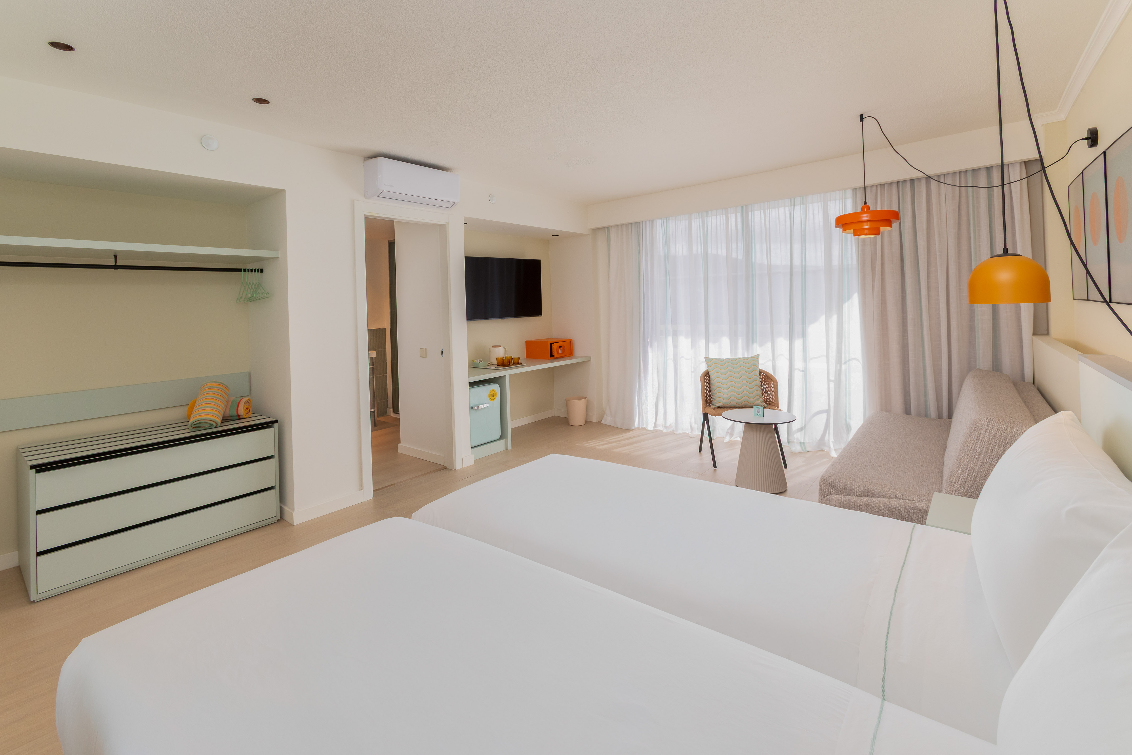 Modern hotel room with two twin beds, bright decor, TV, and seating.