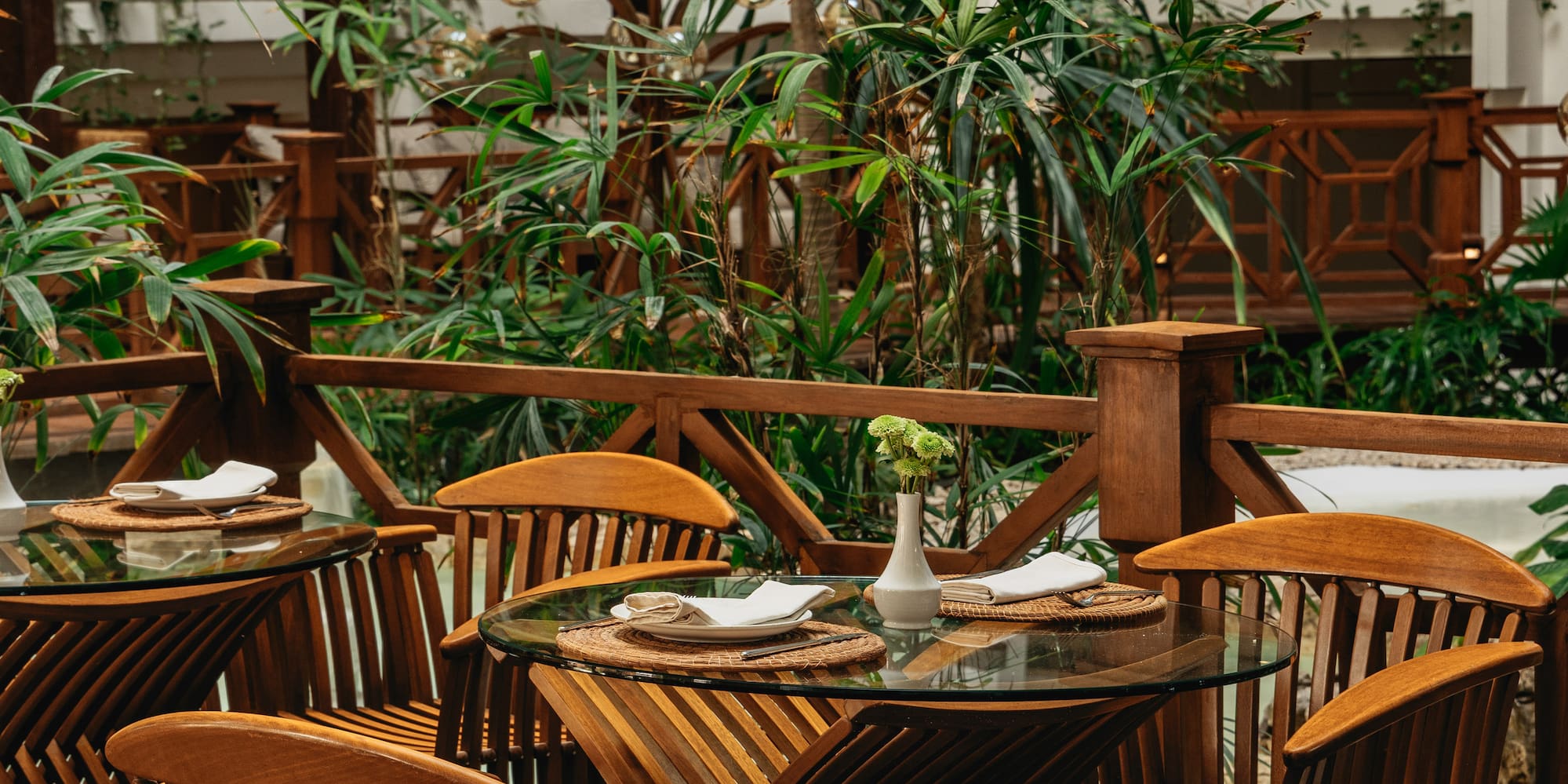 Lush tropical restaurant patio with wooden tables, slatted chairs, and greenery.