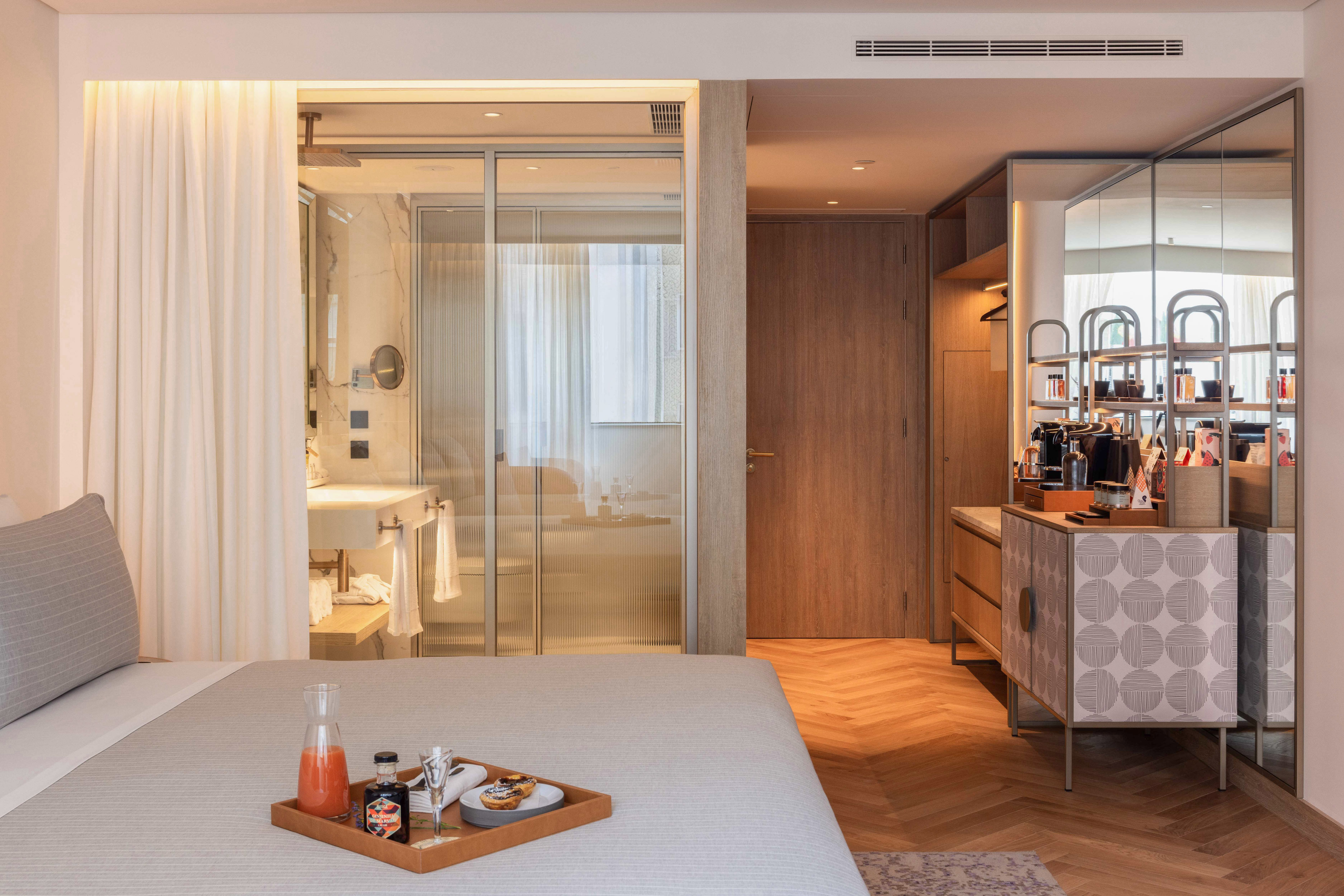 Modern hotel room with breakfast on bed, glass bathroom, and sleek minibar.