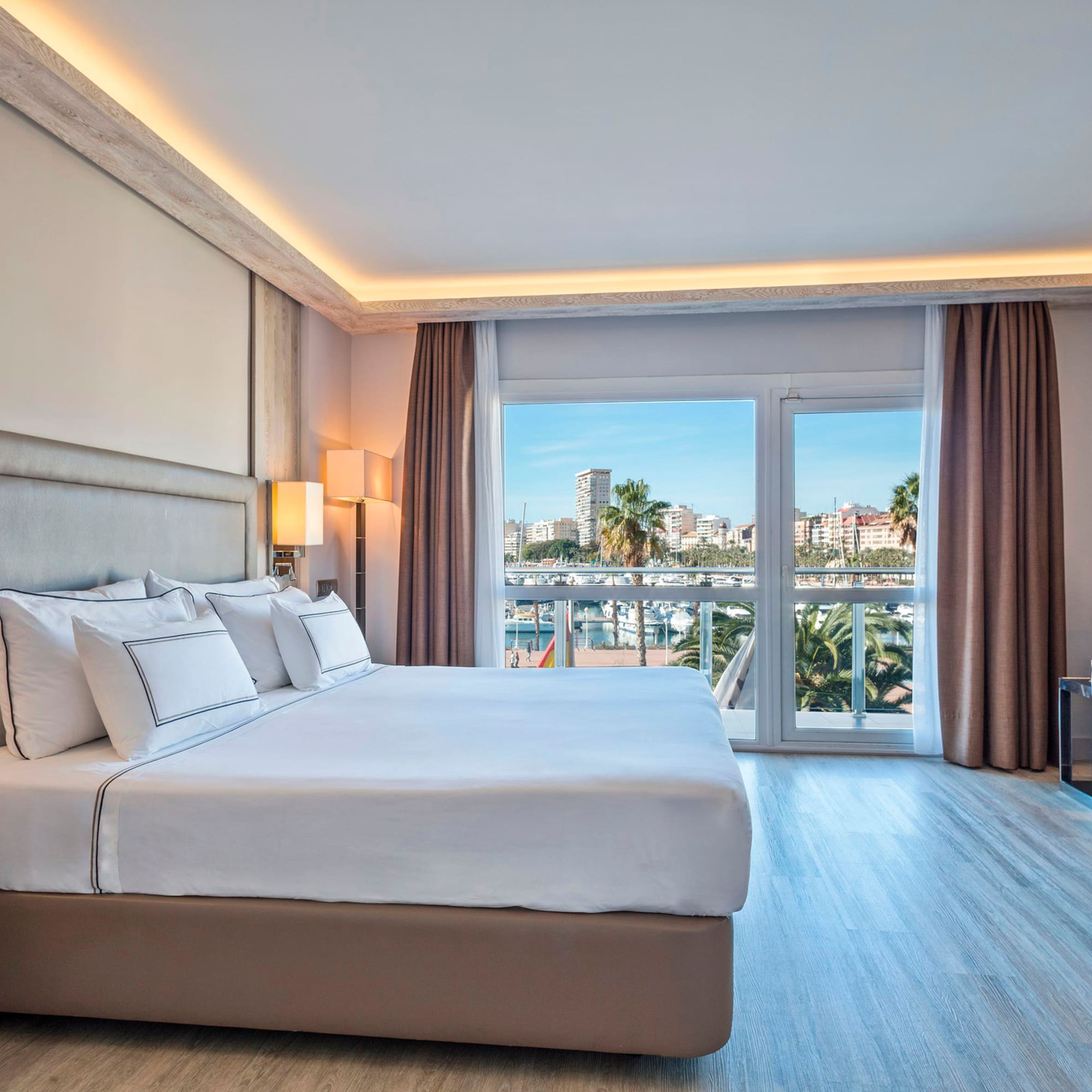 Modern hotel bedroom with large bed and bright marina view from balcony.