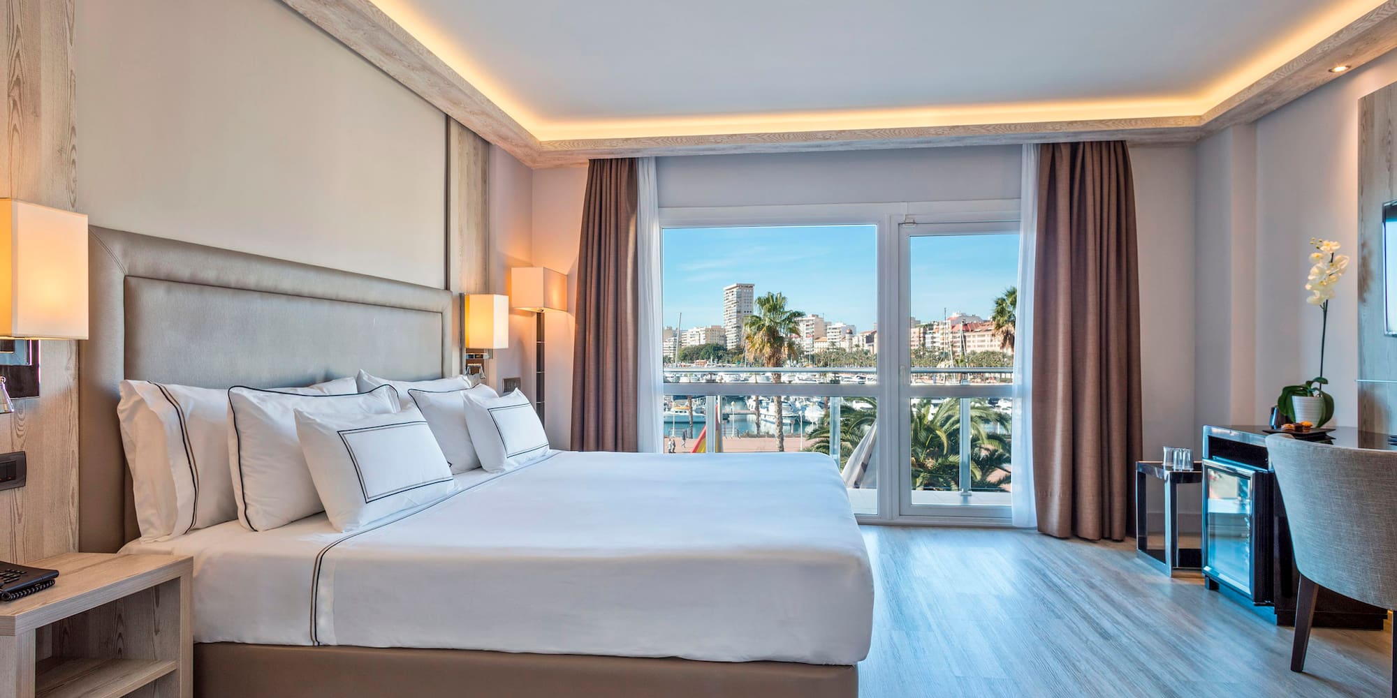 Modern hotel bedroom with large bed and bright marina view from balcony.