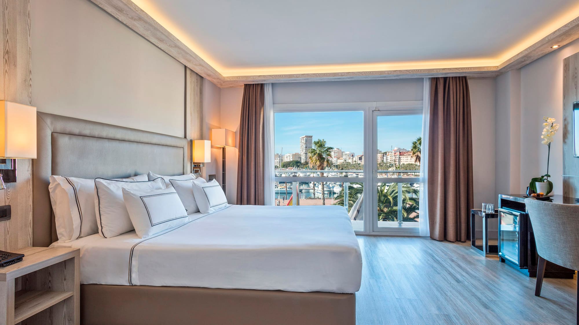 Modern hotel bedroom with large bed and bright marina view from balcony.