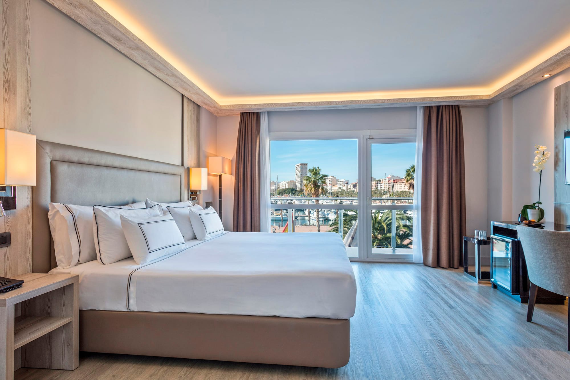 Modern hotel bedroom with large bed and bright marina view from balcony.