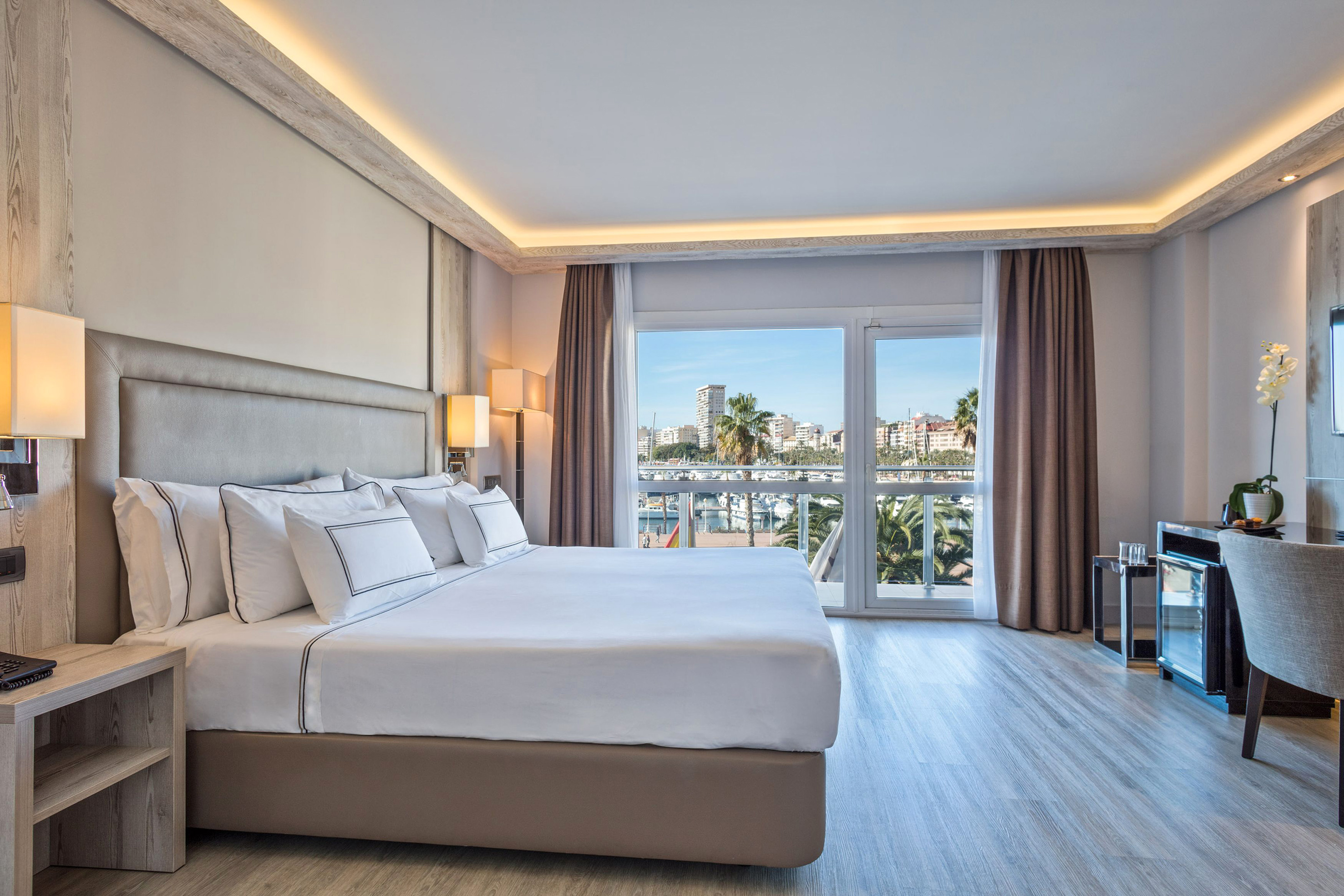 Modern hotel bedroom with large bed and bright marina view from balcony.