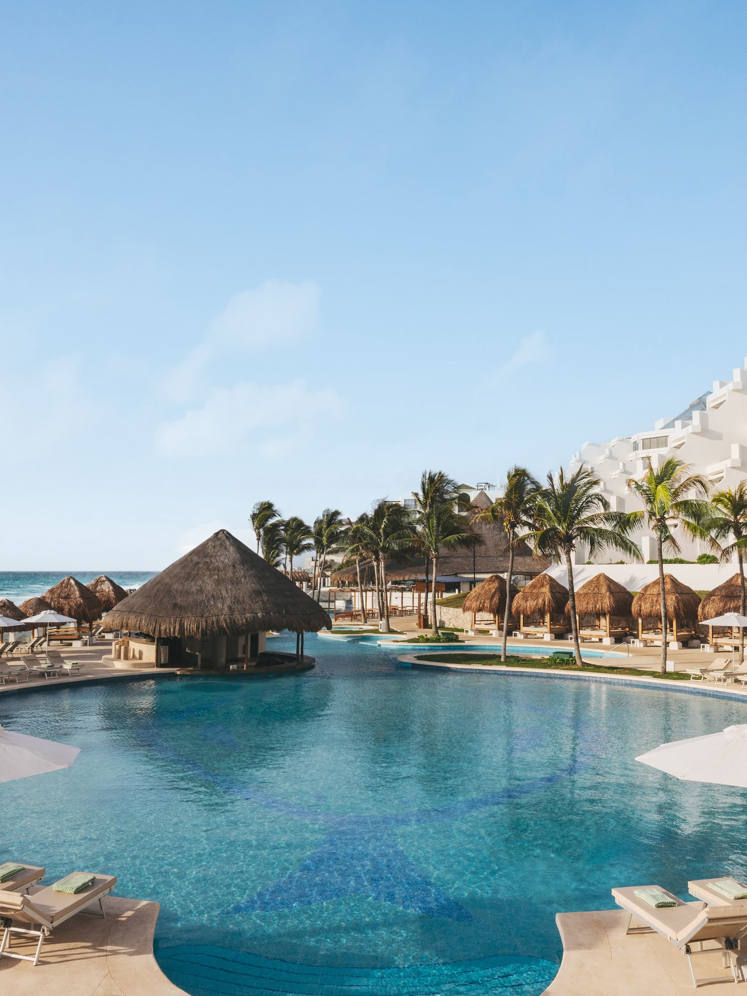 Beach resort pool with swim-up bar, lounge chairs, palm trees, and ocean.