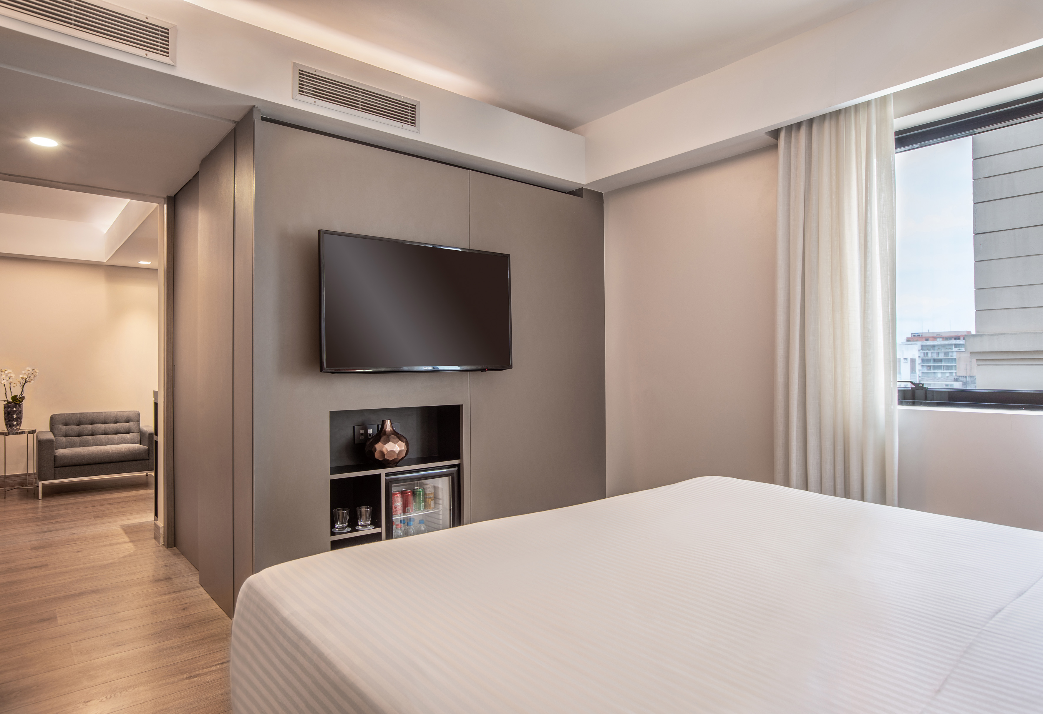 Modern hotel suite bedroom with large bed, wall TV, minibar.