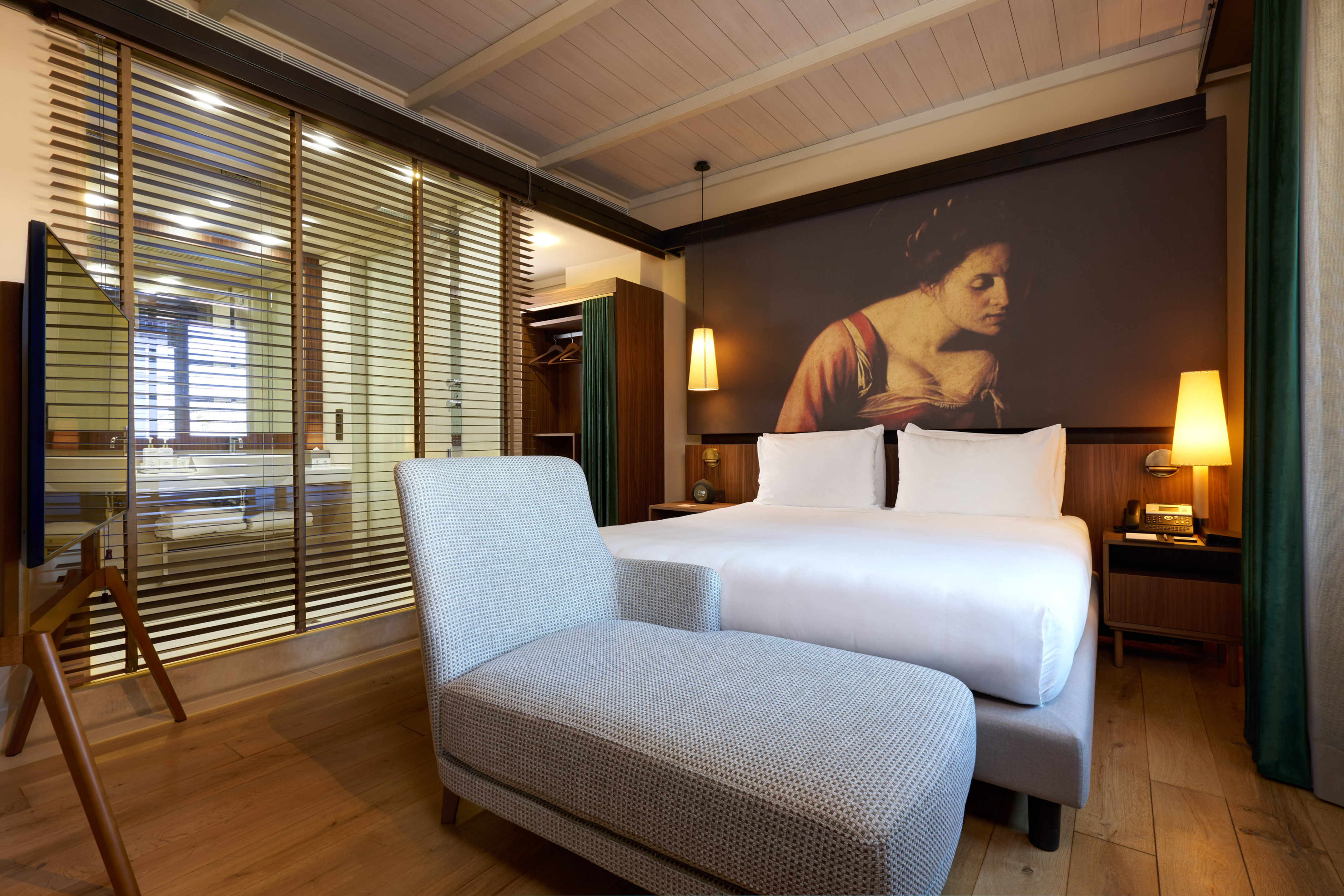 Stylish hotel room with king bed, art mural, chaise lounge, wooden blinds.