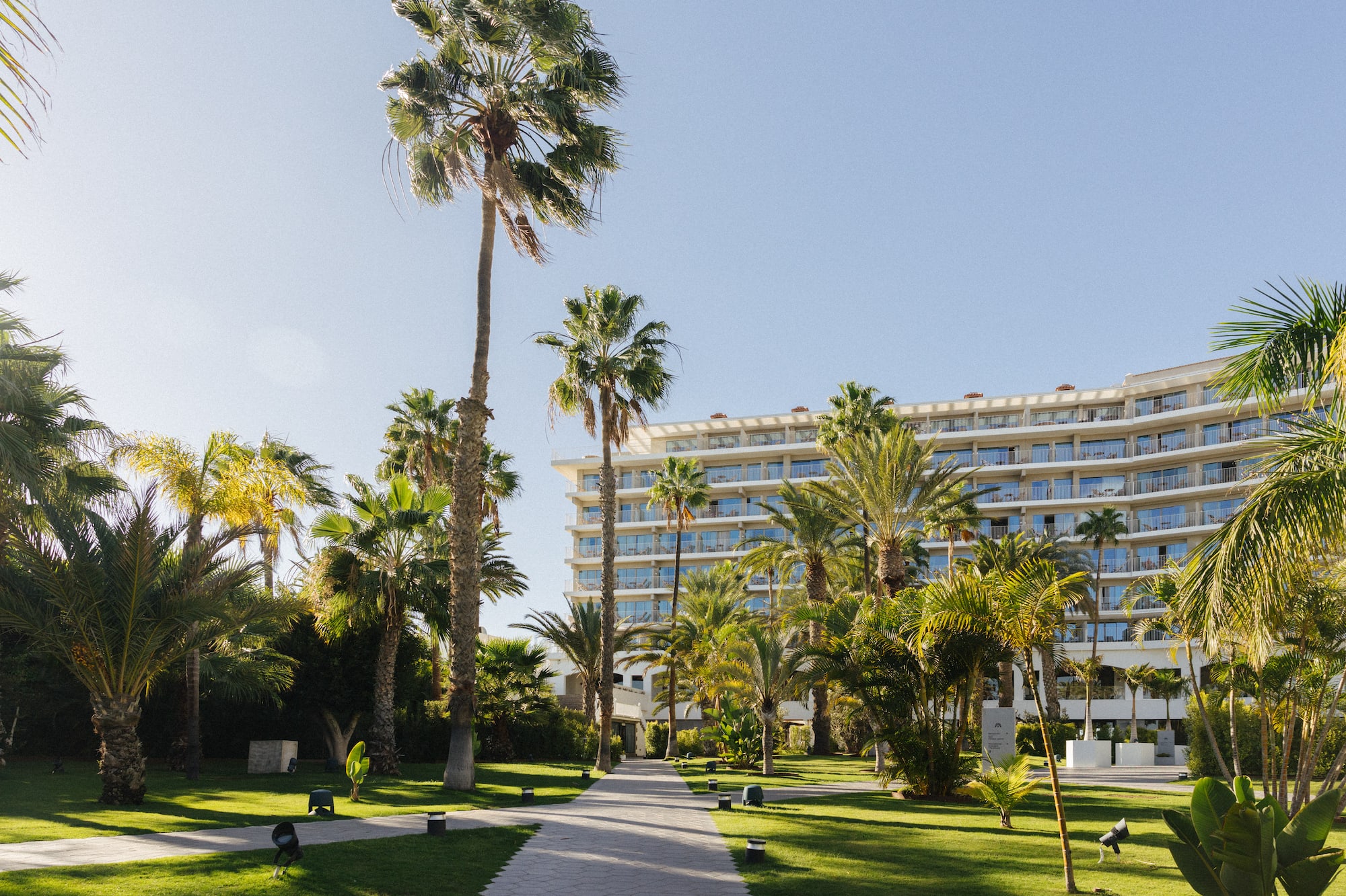 Sunny resort hotel with palm trees, green lawns, and a paved pathway.