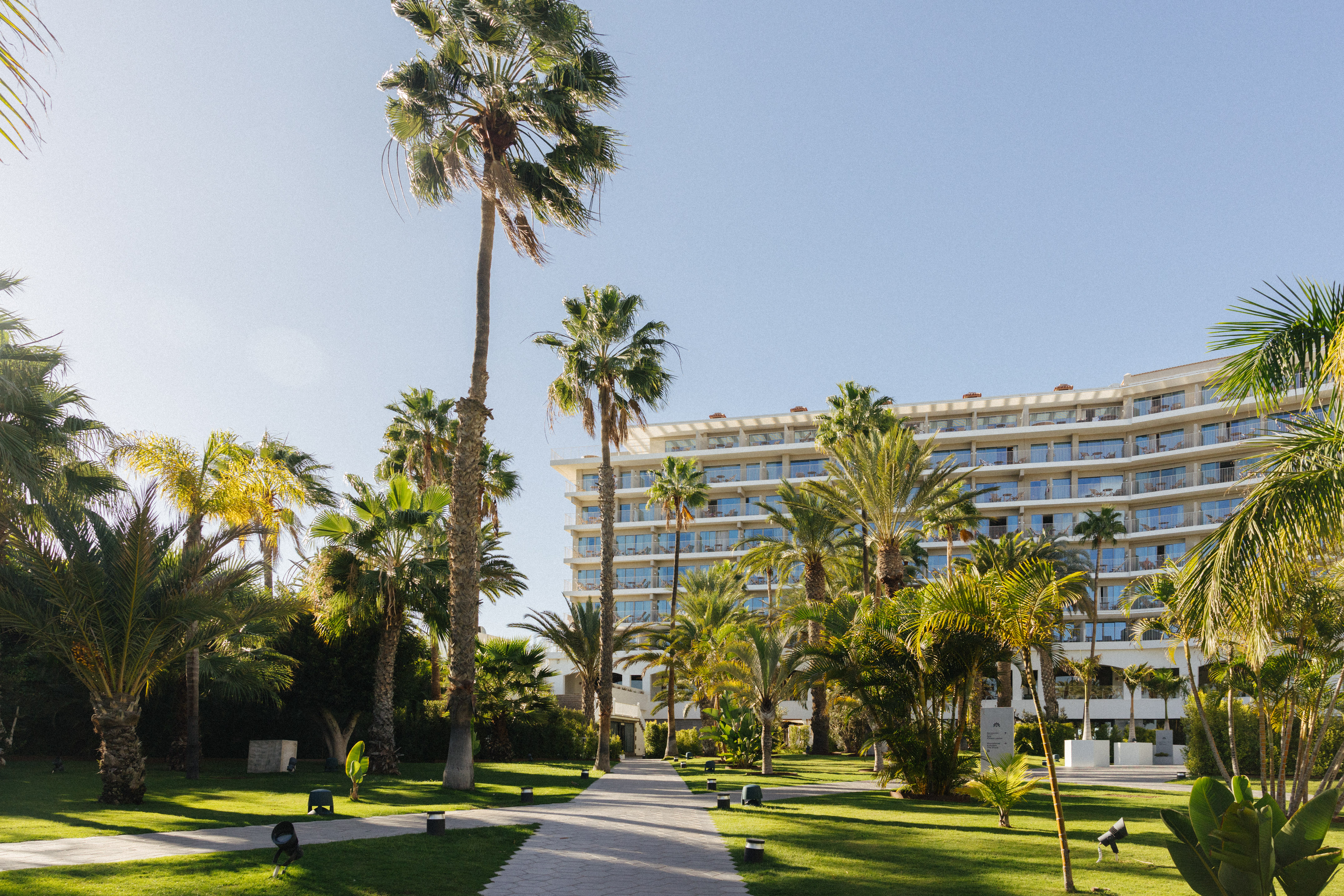 Sunny resort hotel with palm trees, green lawns, and a paved pathway.