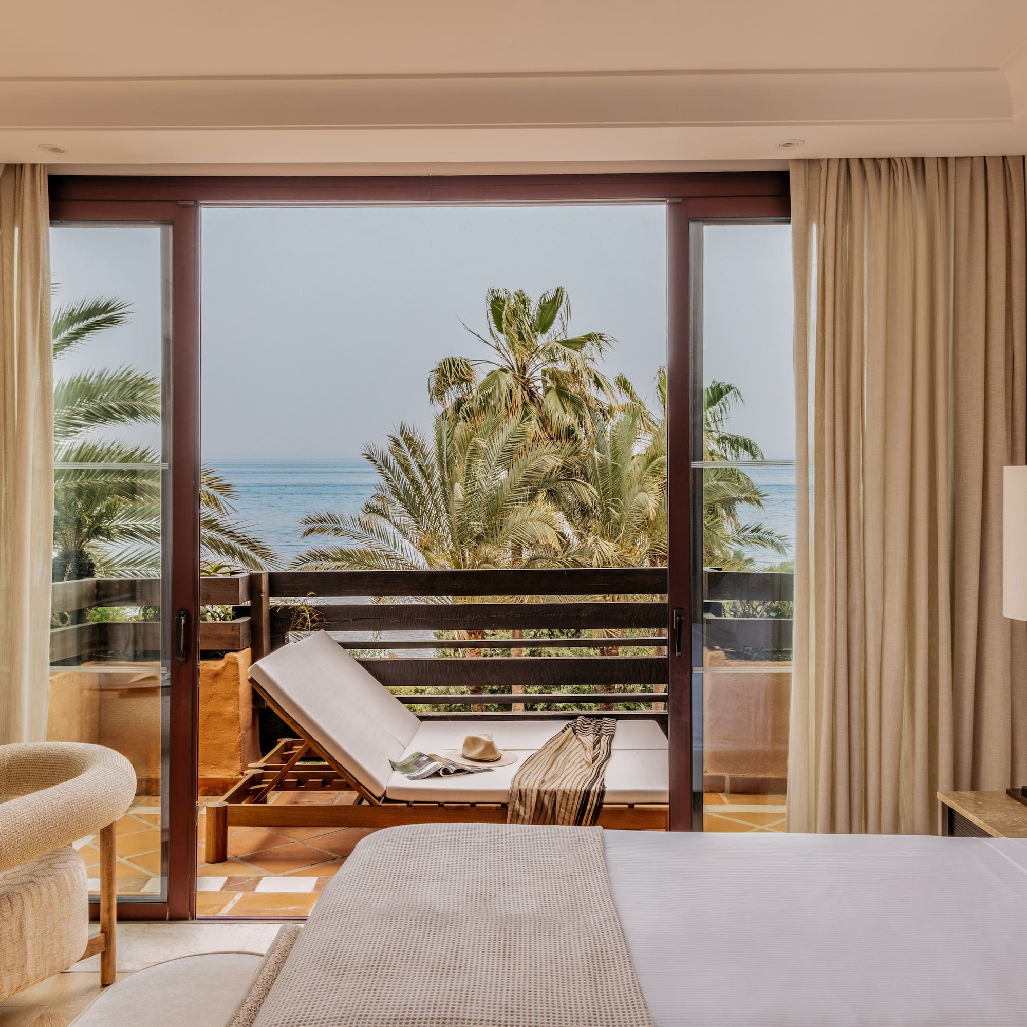 Hotel room with ocean view balcony, palm trees, and lounge chair.