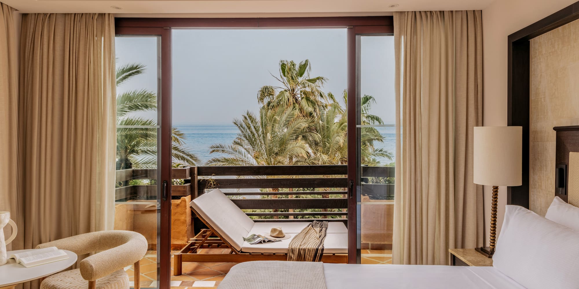 Hotel room with ocean view balcony, palm trees, and lounge chair.