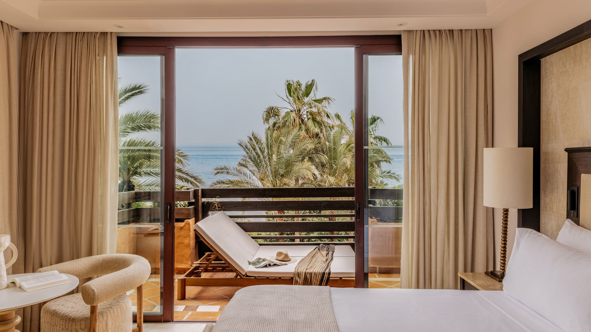 Hotel room with ocean view balcony, palm trees, and lounge chair.