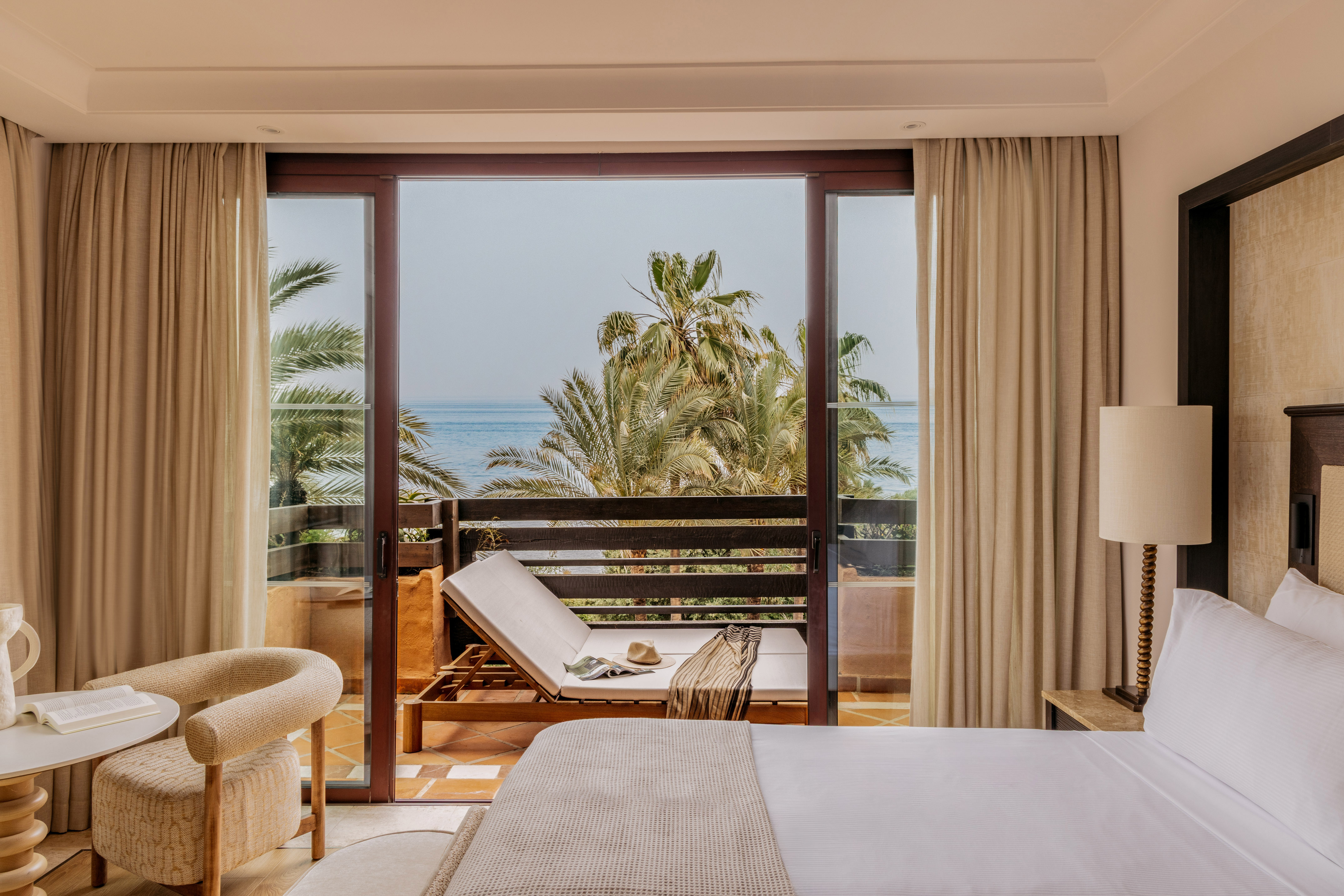 Hotel room with ocean view balcony, palm trees, and lounge chair.
