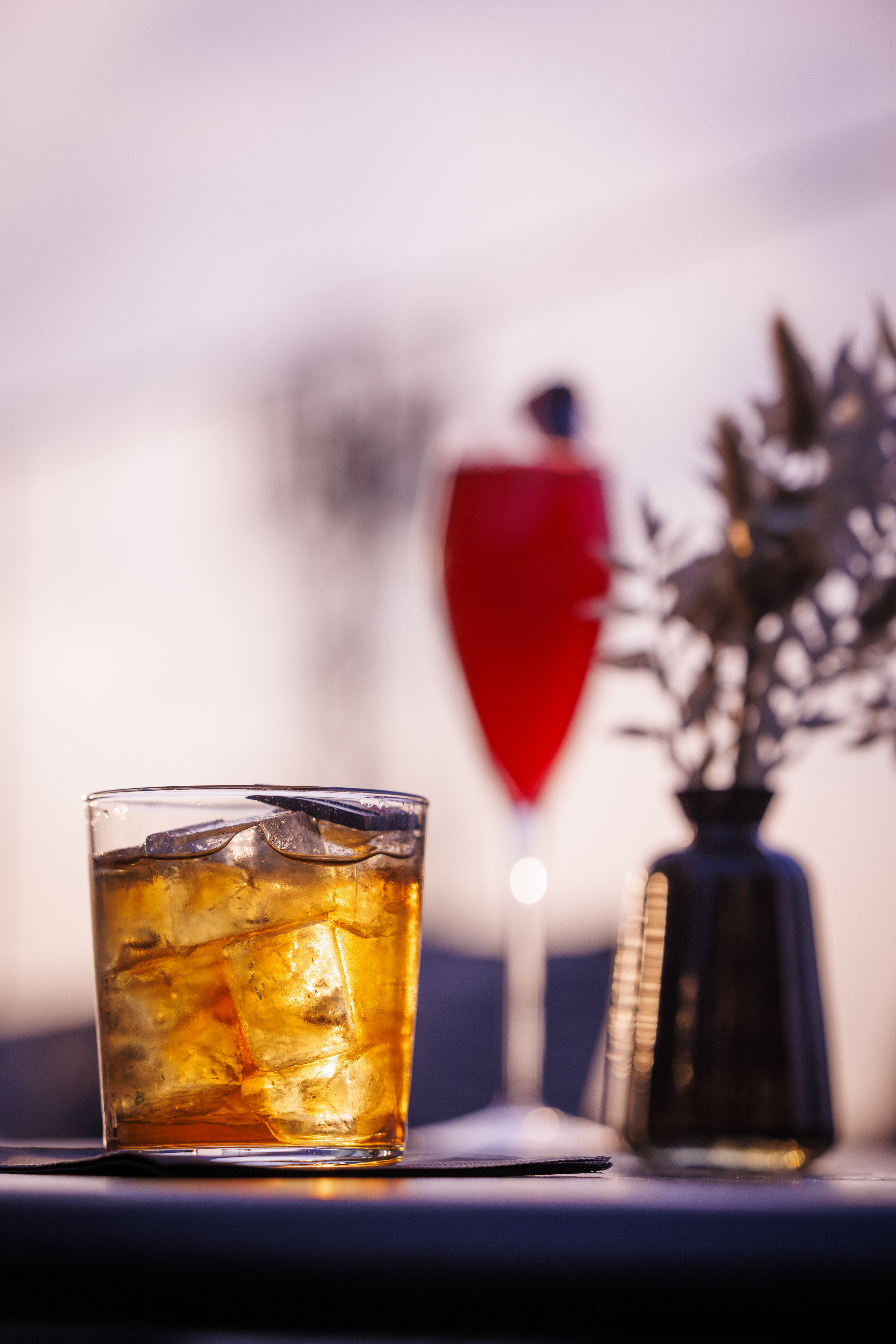 Iced golden cocktail, red drink, and vase on a bar, ambient evening light.