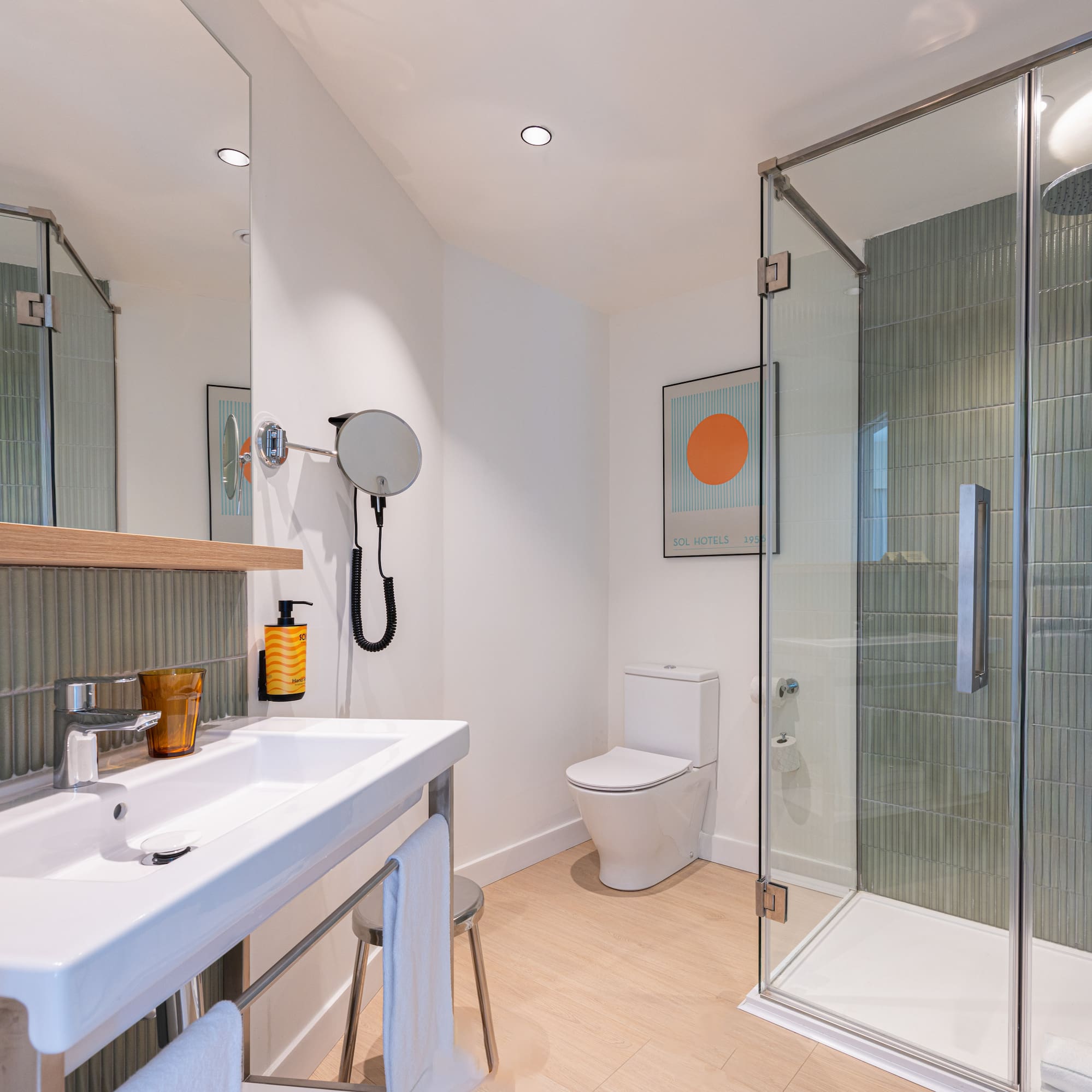 Modern hotel bathroom with white sink, toilet, glass shower, green tiles, and wood floor.