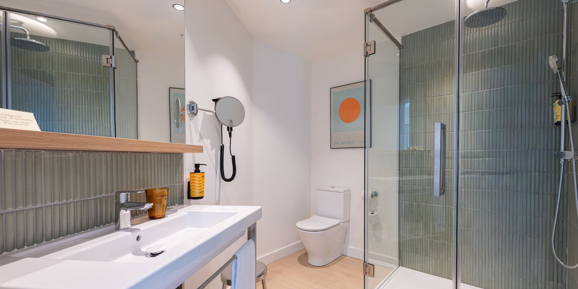 Modern hotel bathroom with white sink, toilet, glass shower, green tiles, and wood floor.