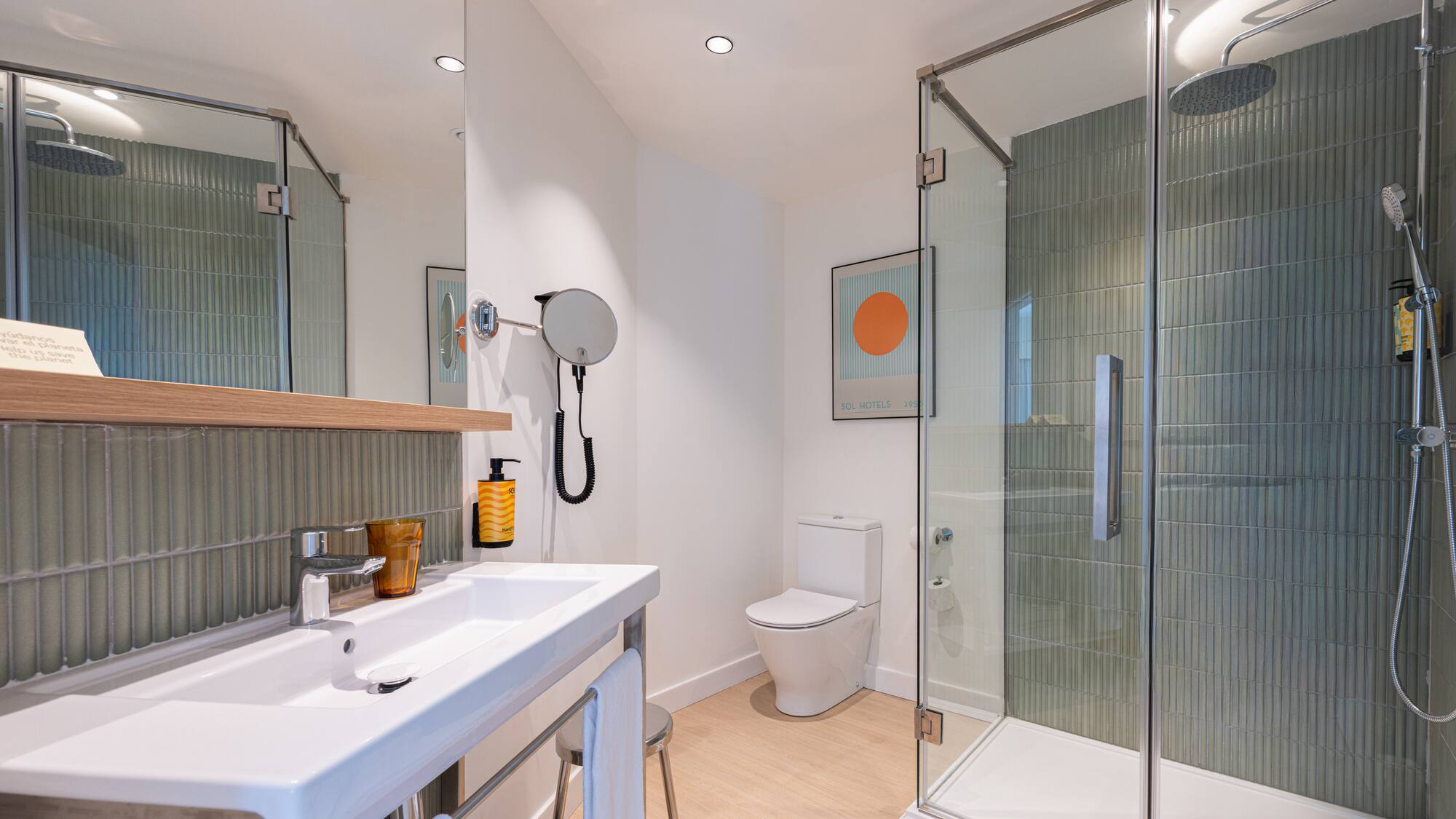 Modern hotel bathroom with white sink, toilet, glass shower, green tiles, and wood floor.