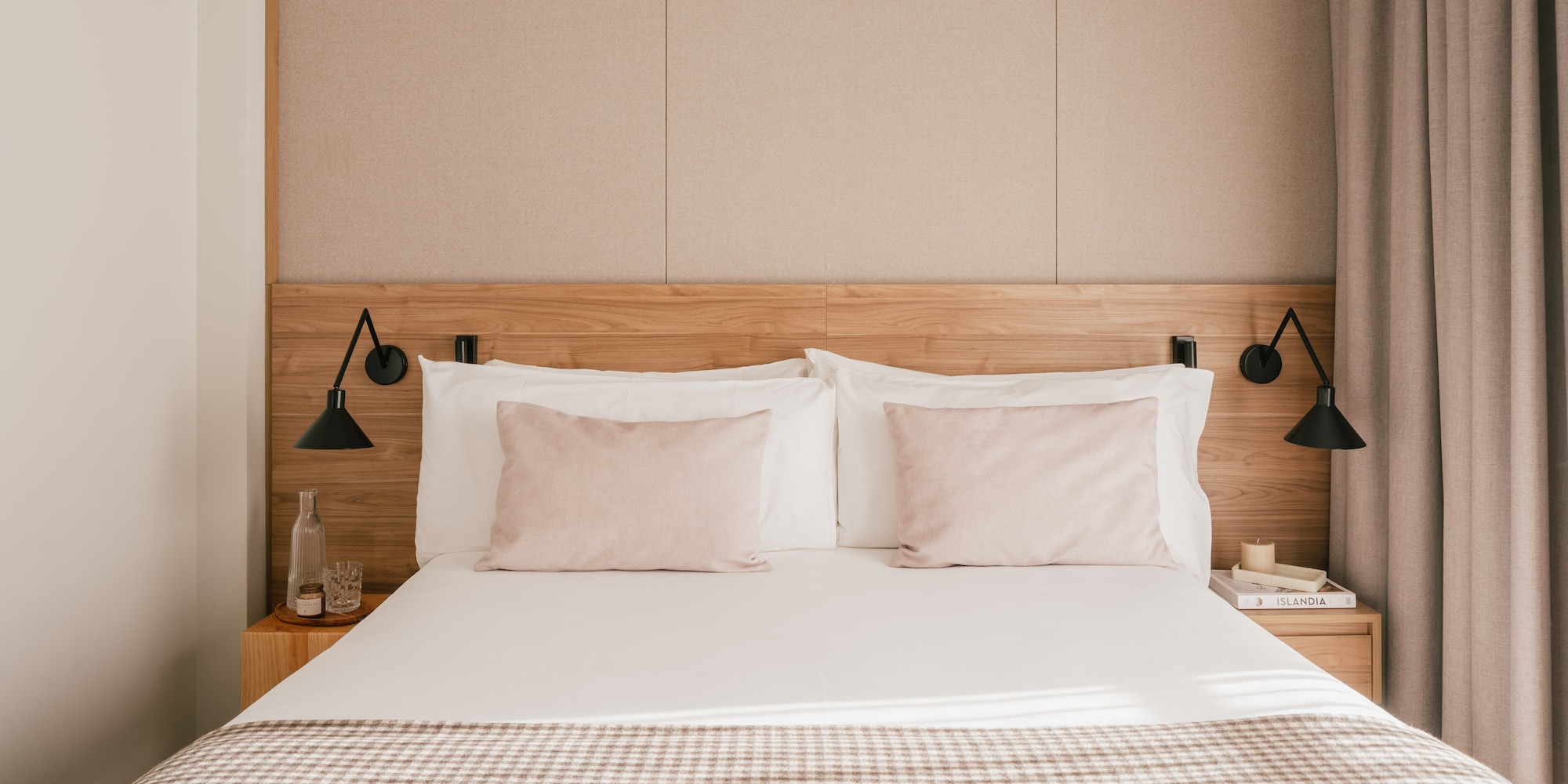 Modern hotel bedroom with wooden headboard, pink pillows, and plaid throw blanket.