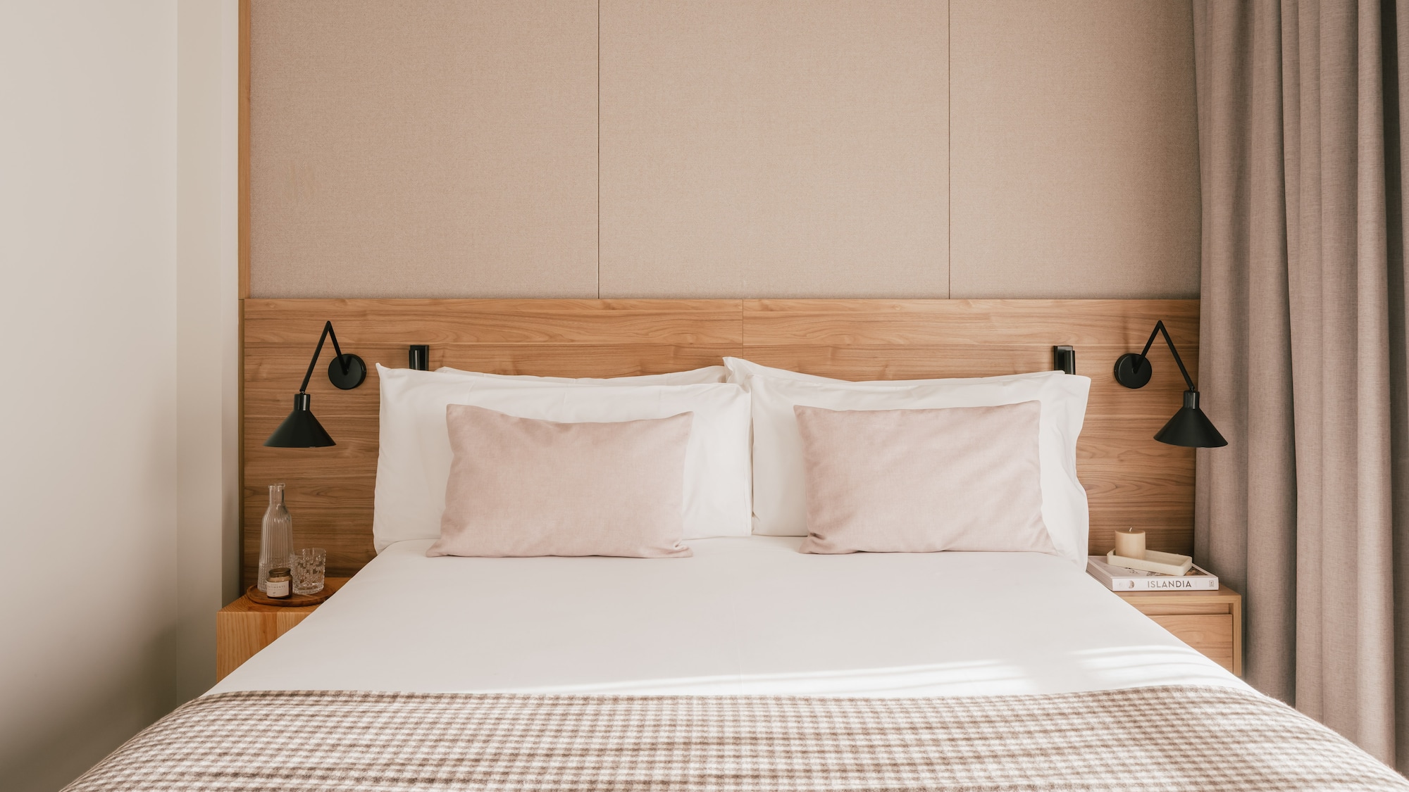 Modern hotel bedroom with wooden headboard, pink pillows, and plaid throw blanket.