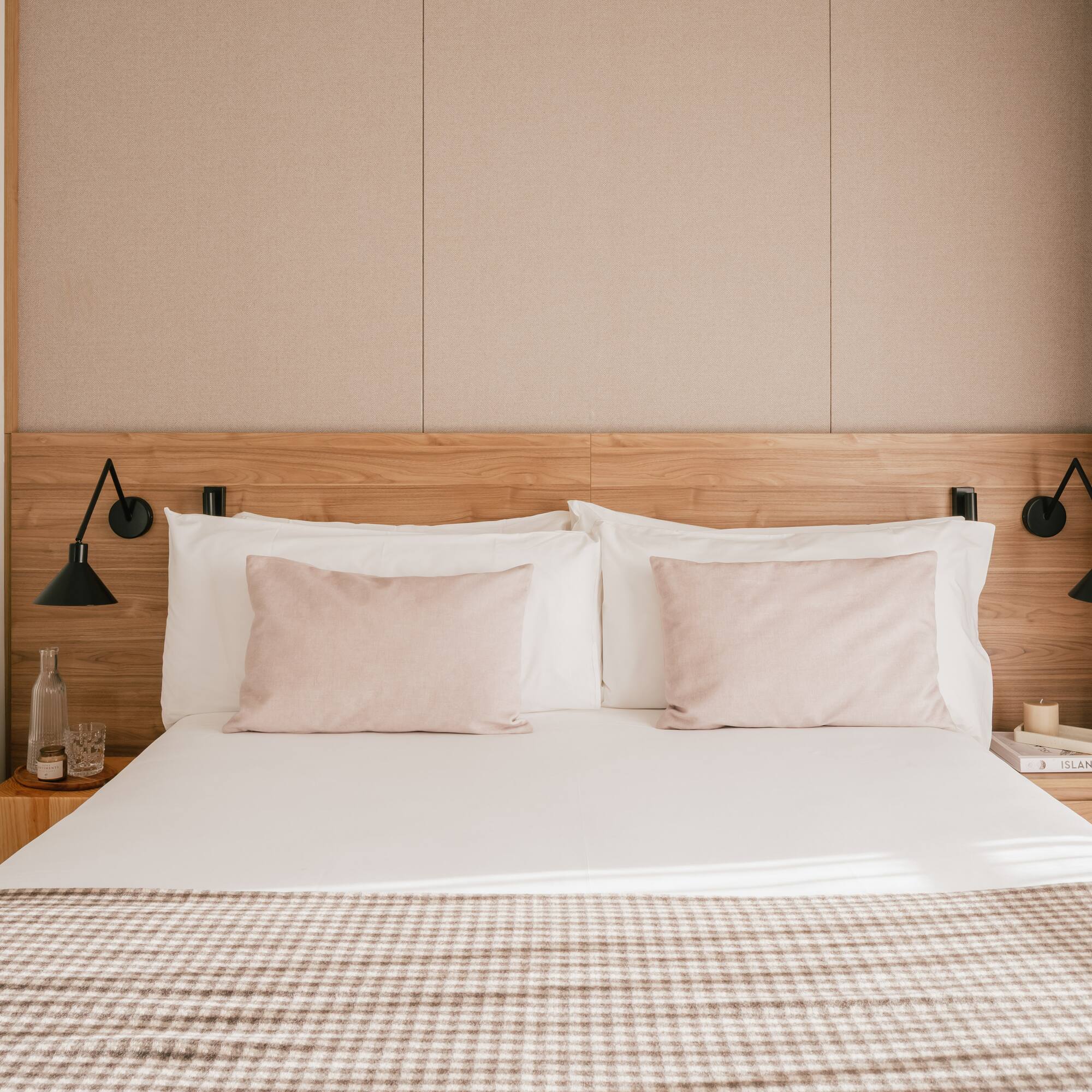 Modern hotel bedroom with wooden headboard, pink pillows, and plaid throw blanket.