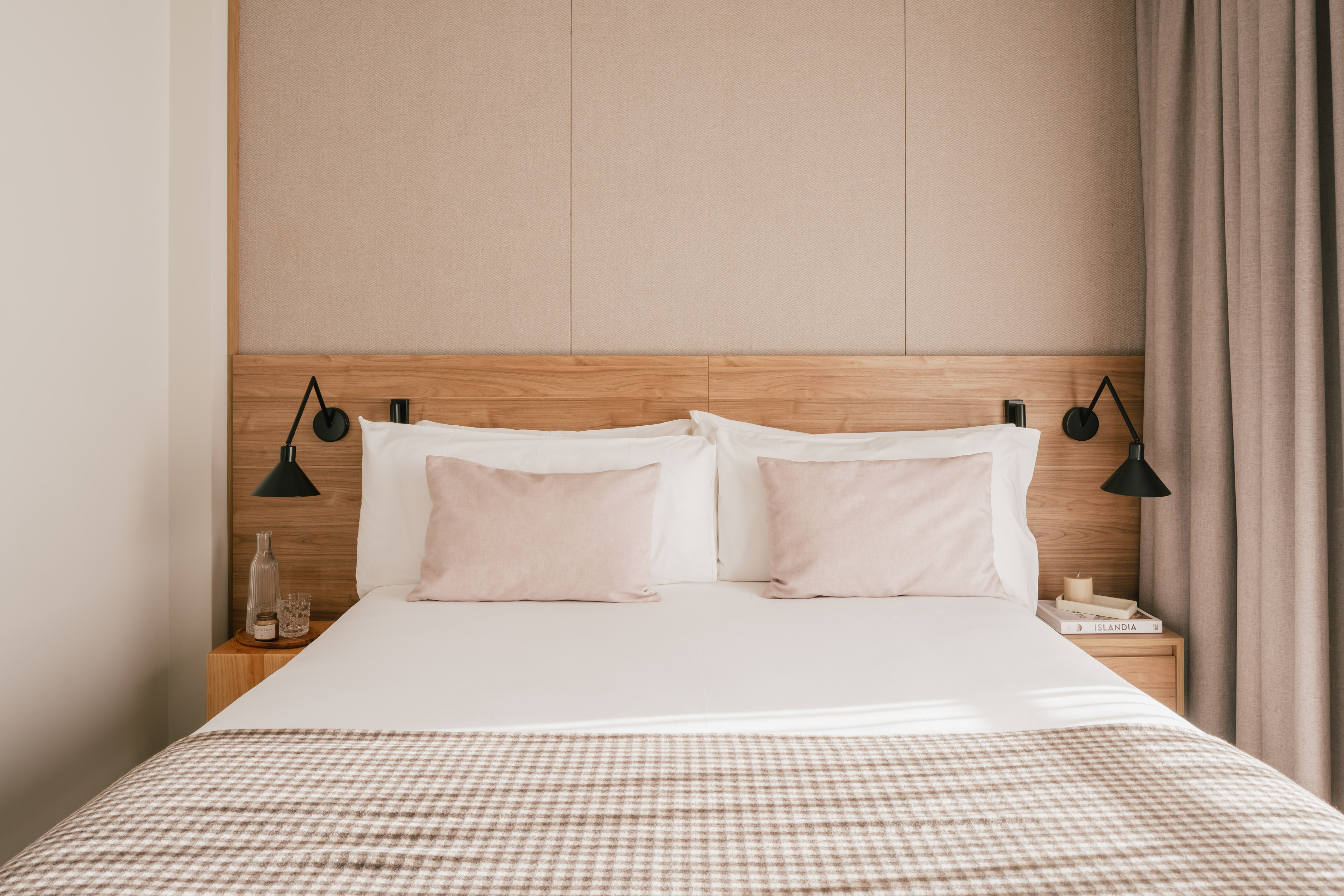 Modern hotel bedroom with wooden headboard, pink pillows, and plaid throw blanket.