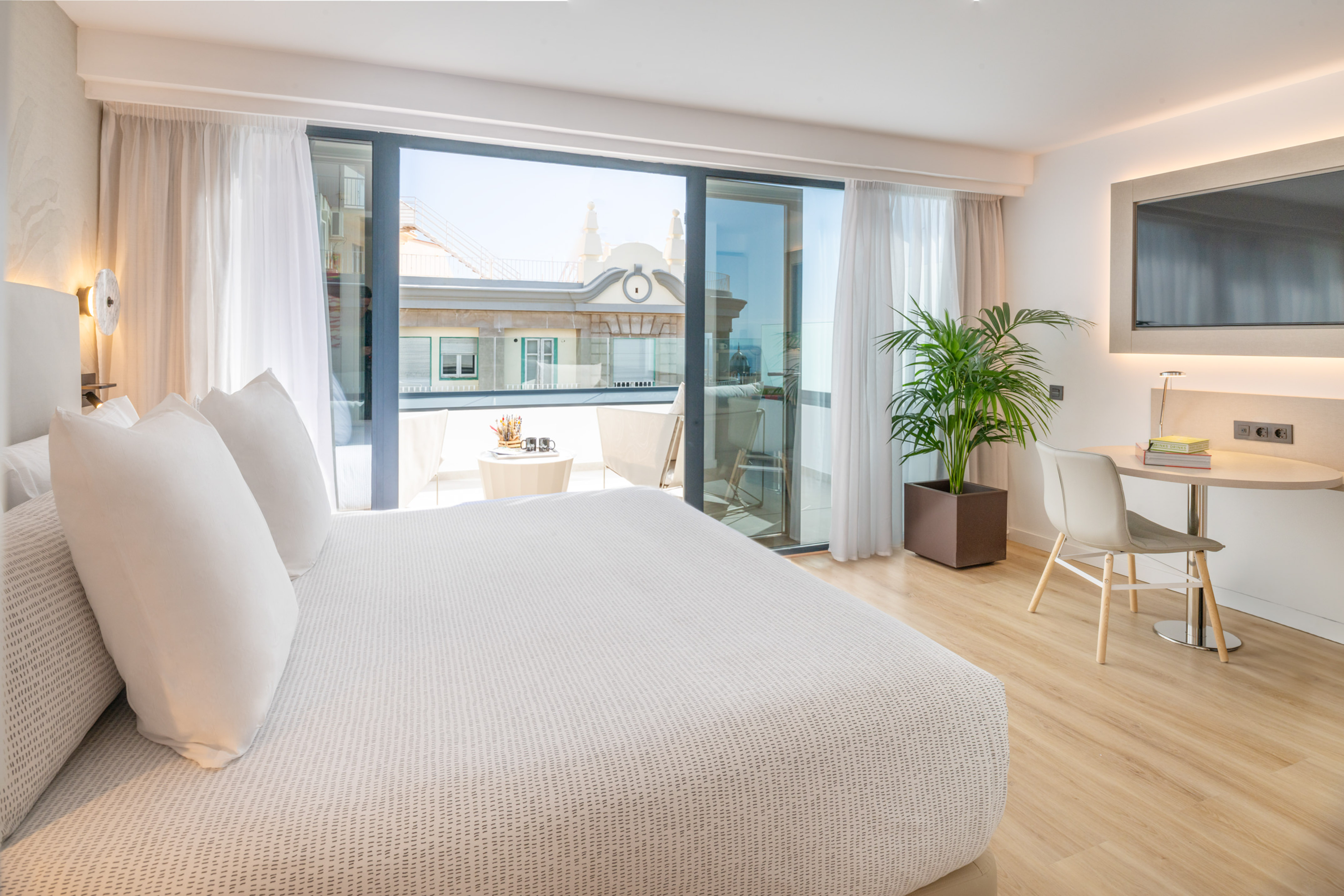 Modern hotel room with white bed, work desk, and balcony city view.