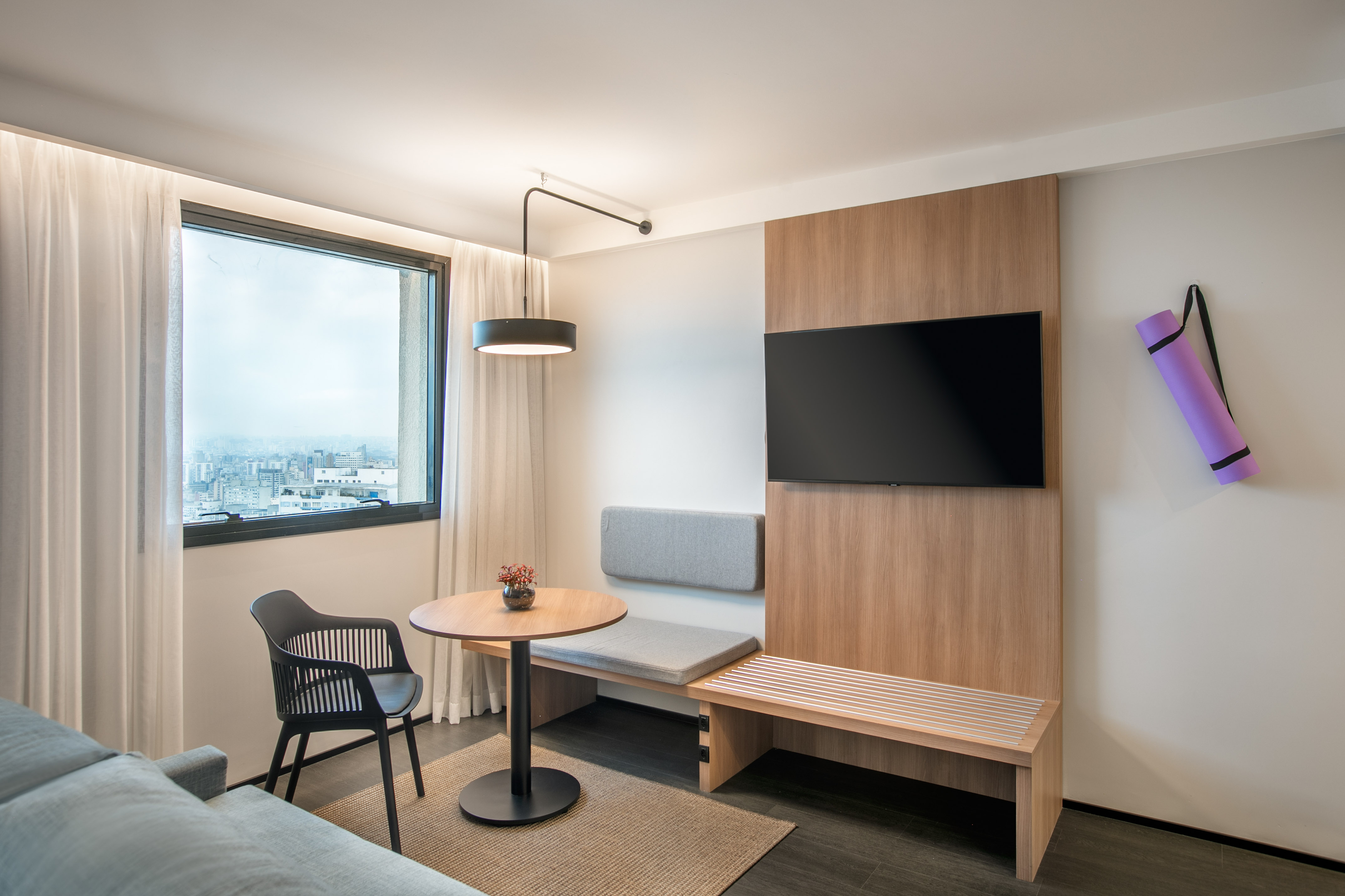 Modern hotel room with city view, TV, small table, bench, and purple yoga mat.
