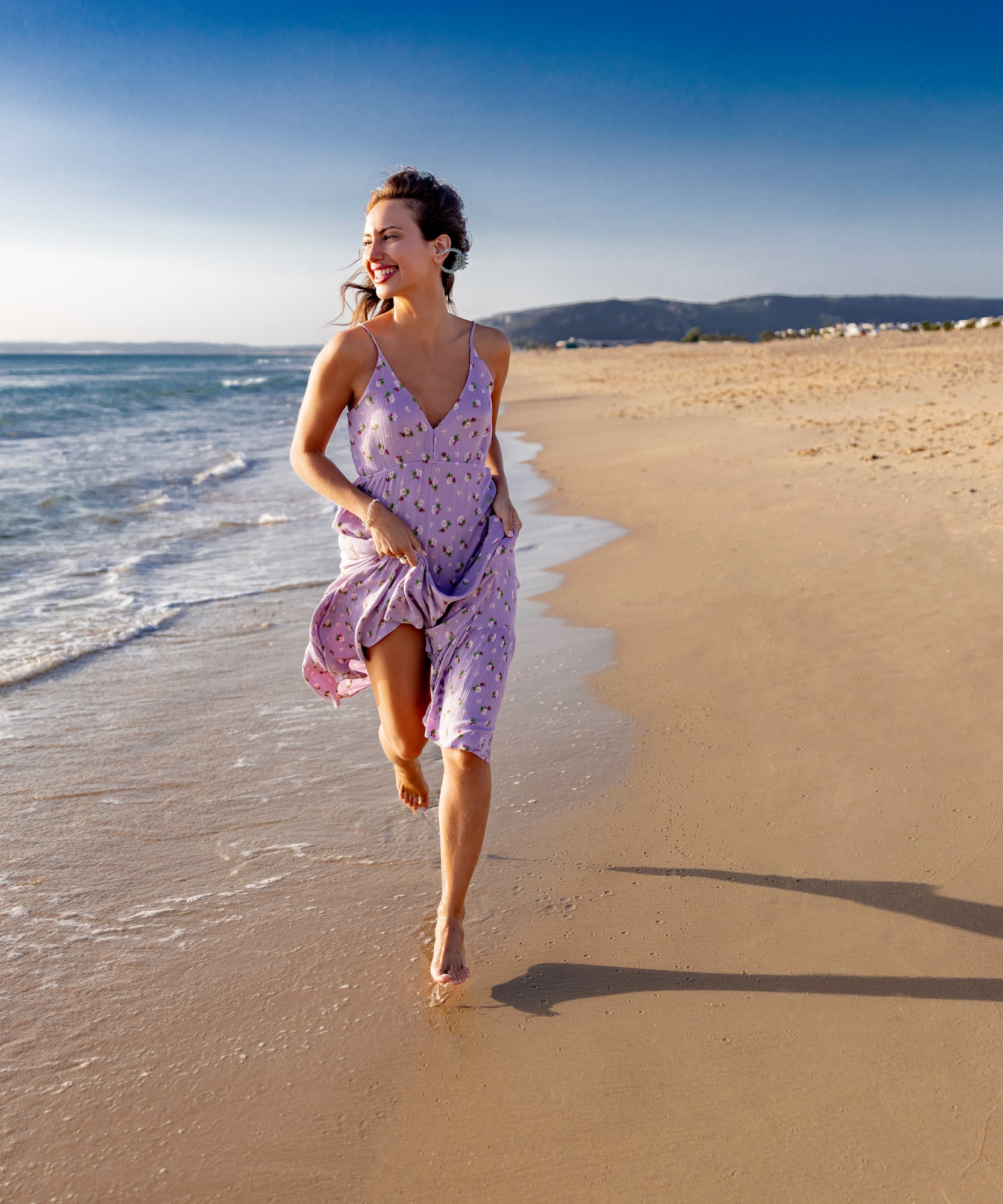 Woman in purple summer dress running barefoot on a sunny beach.