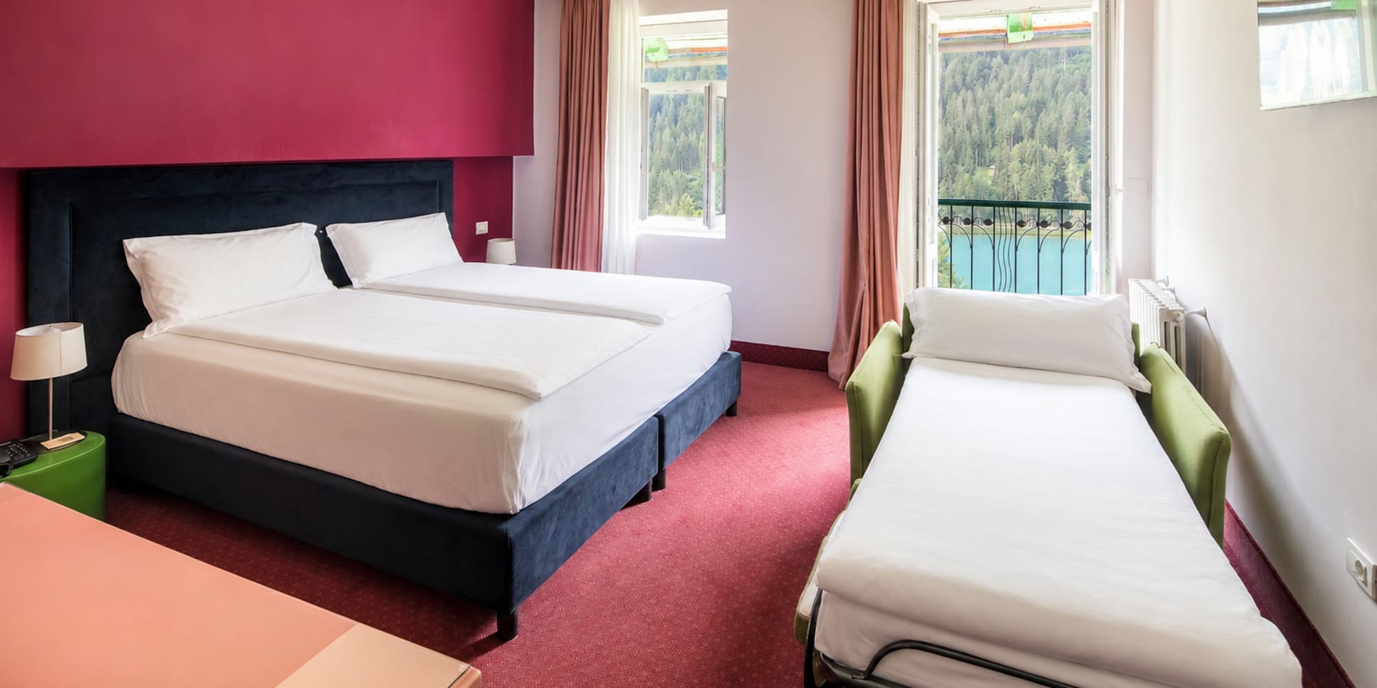 Hotel room with twin beds, sofa bed, red decor, and balcony lake view.