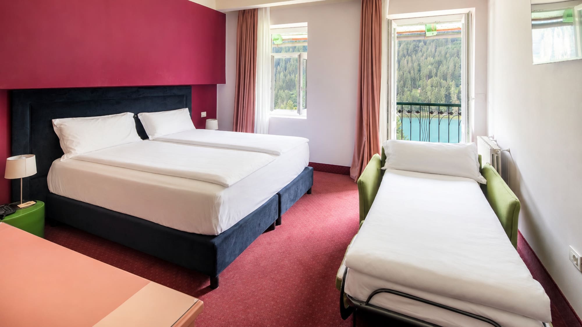 Hotel room with twin beds, sofa bed, red decor, and balcony lake view.