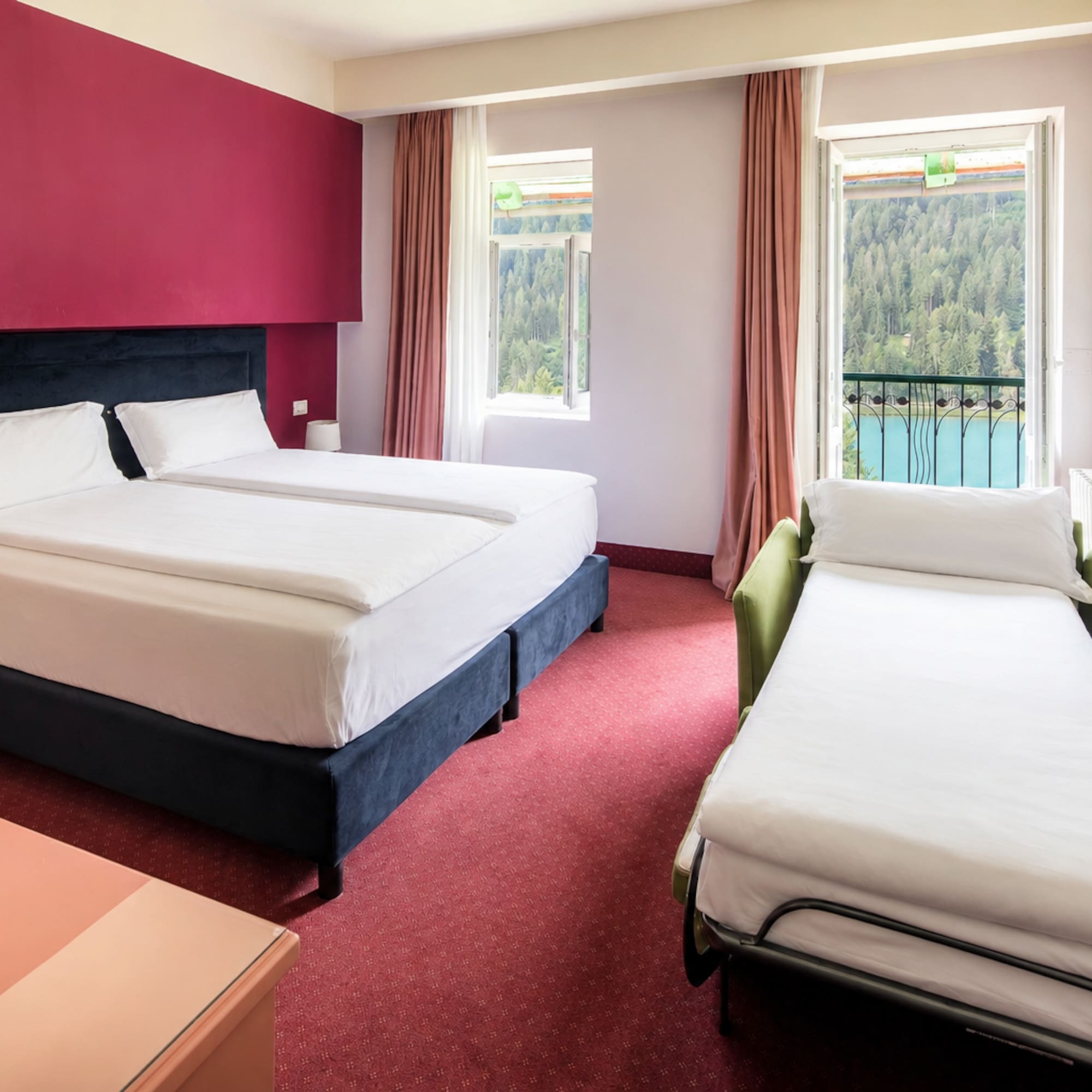 Hotel room with twin beds, sofa bed, red decor, and balcony lake view.