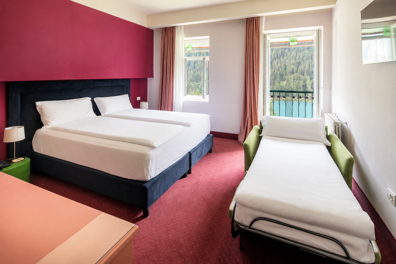 Hotel room with twin beds, sofa bed, red decor, and balcony lake view.
